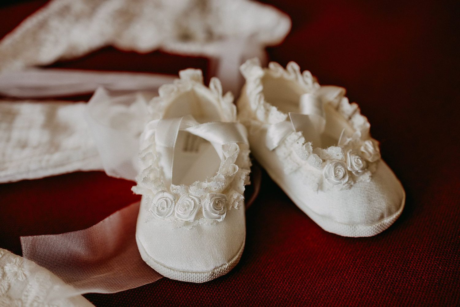 Delicate white baby shoes with rose details on red background for christening ceremony.