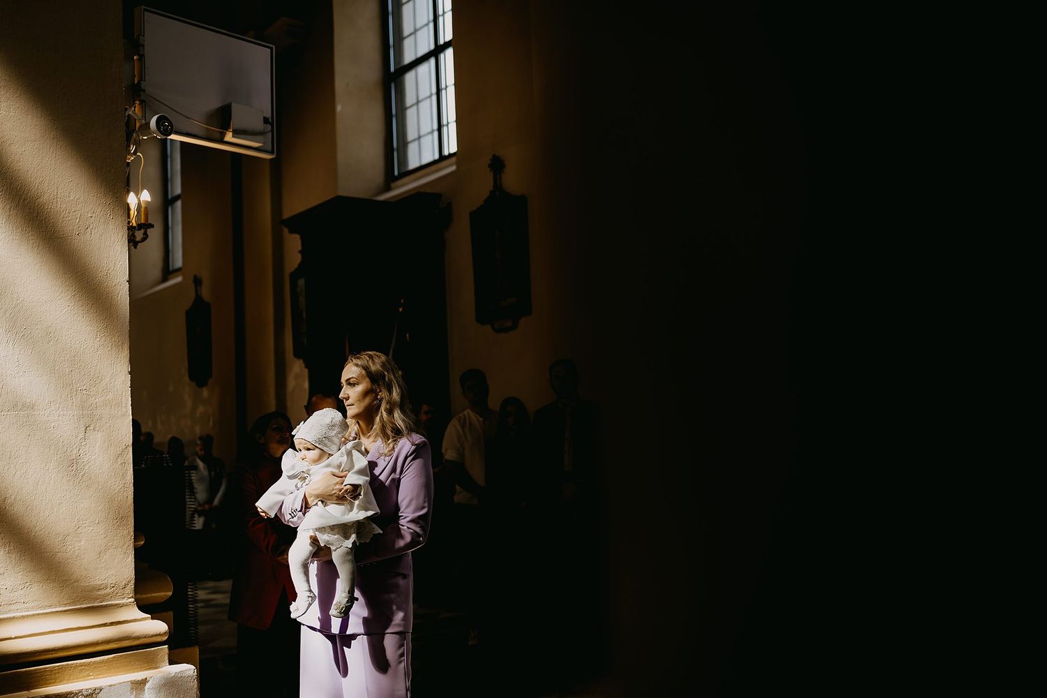 Woman in lavender dress holds infant in white christening gown inside historic church.