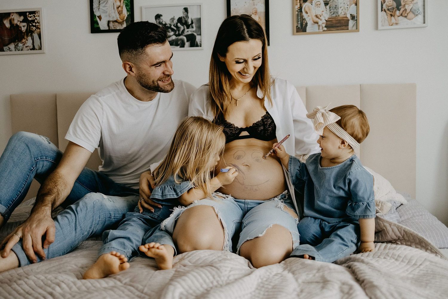 Family sitting on bed with pregnant mother as two young children touch her baby bump.