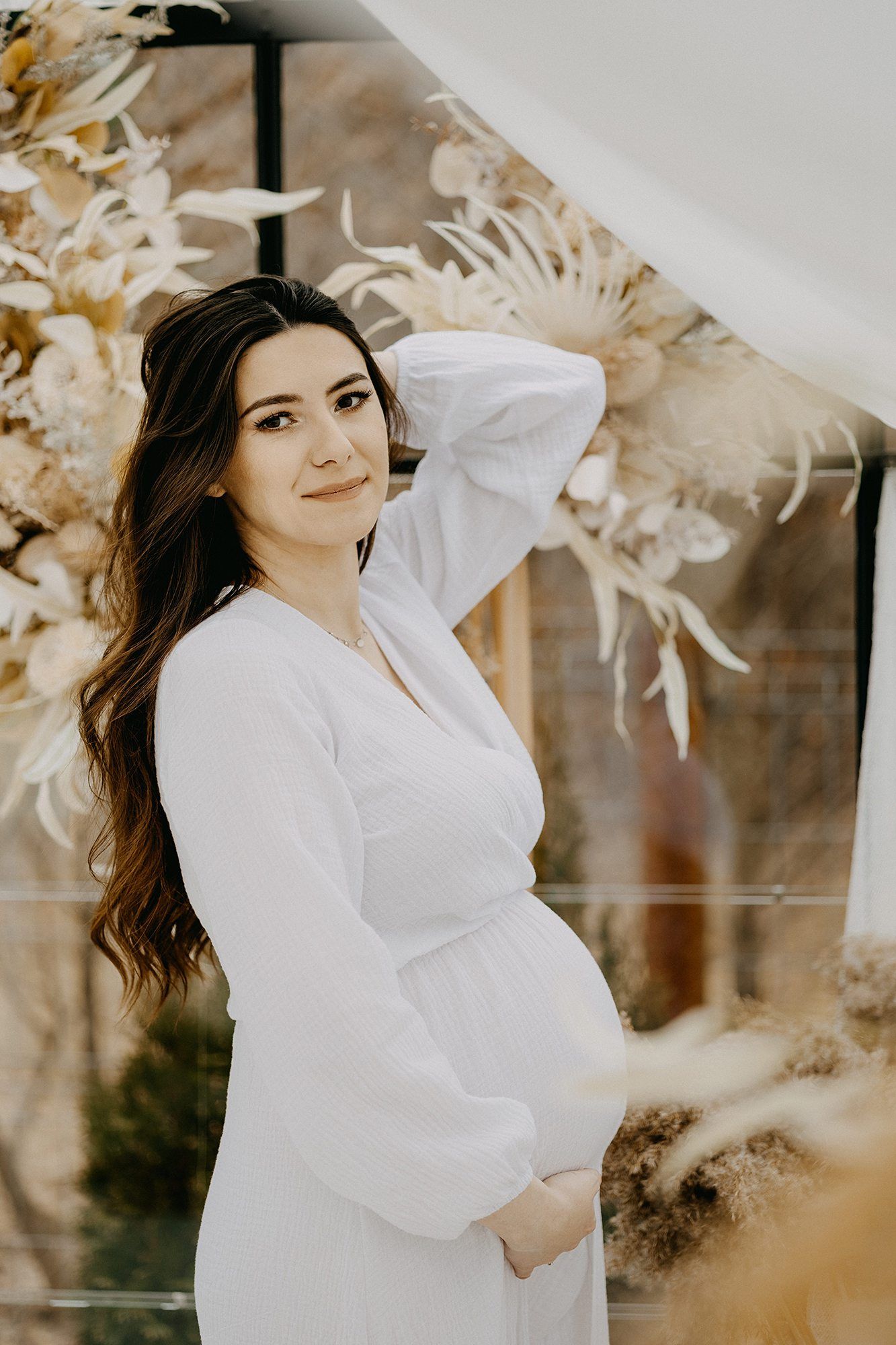 Elegant pregnant woman in white dress posing with hand in hair before boho floral backdrop.