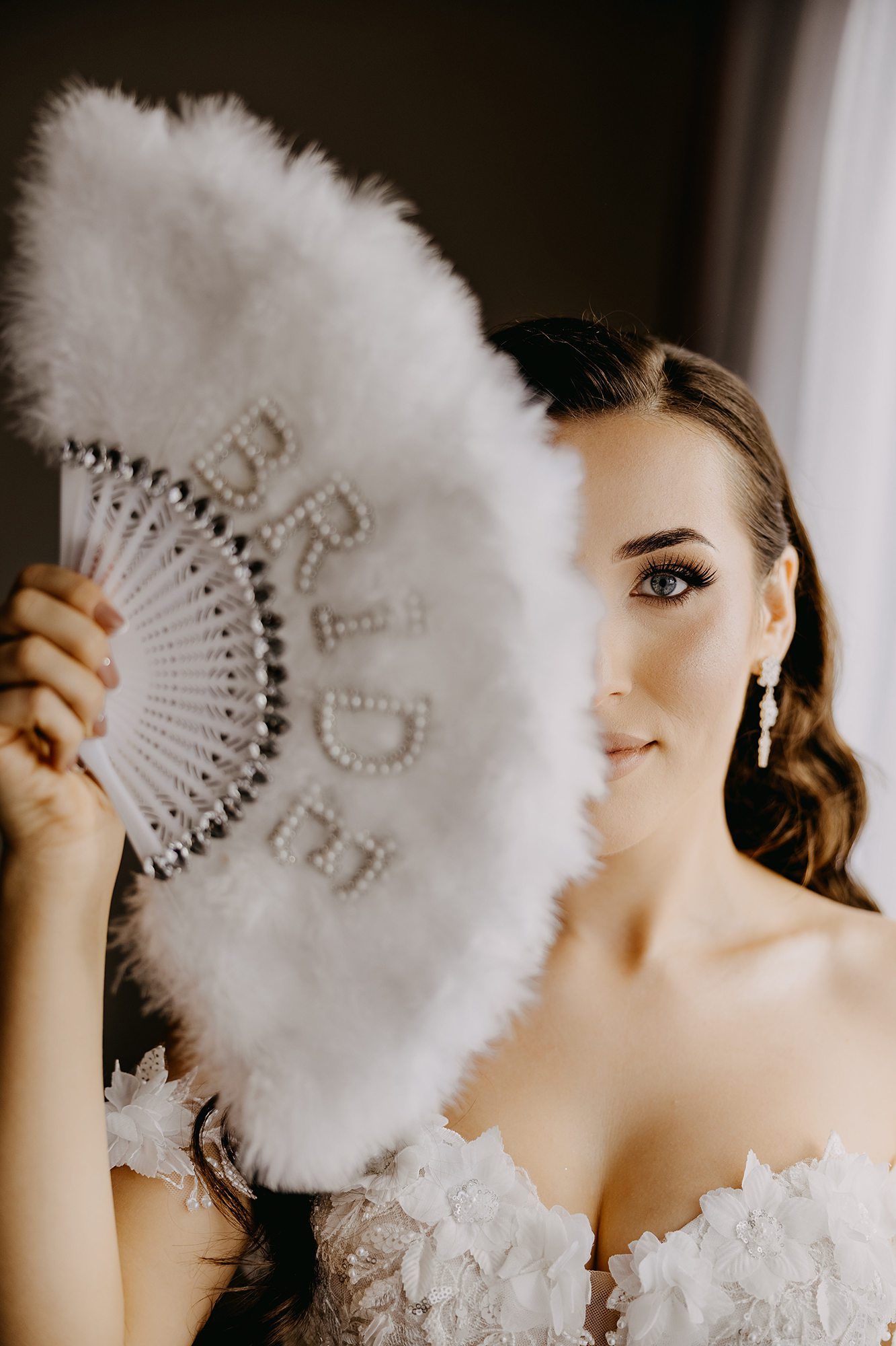 Bride holding white feather fan with pearl BRIDE lettering covering half her face.