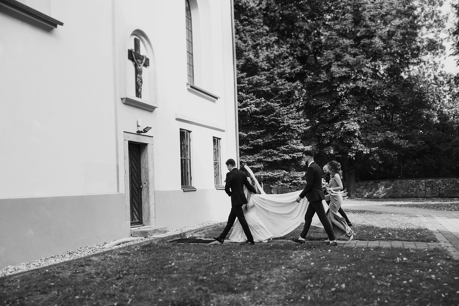 Black and white photo of wedding party carrying bridal train outside church.