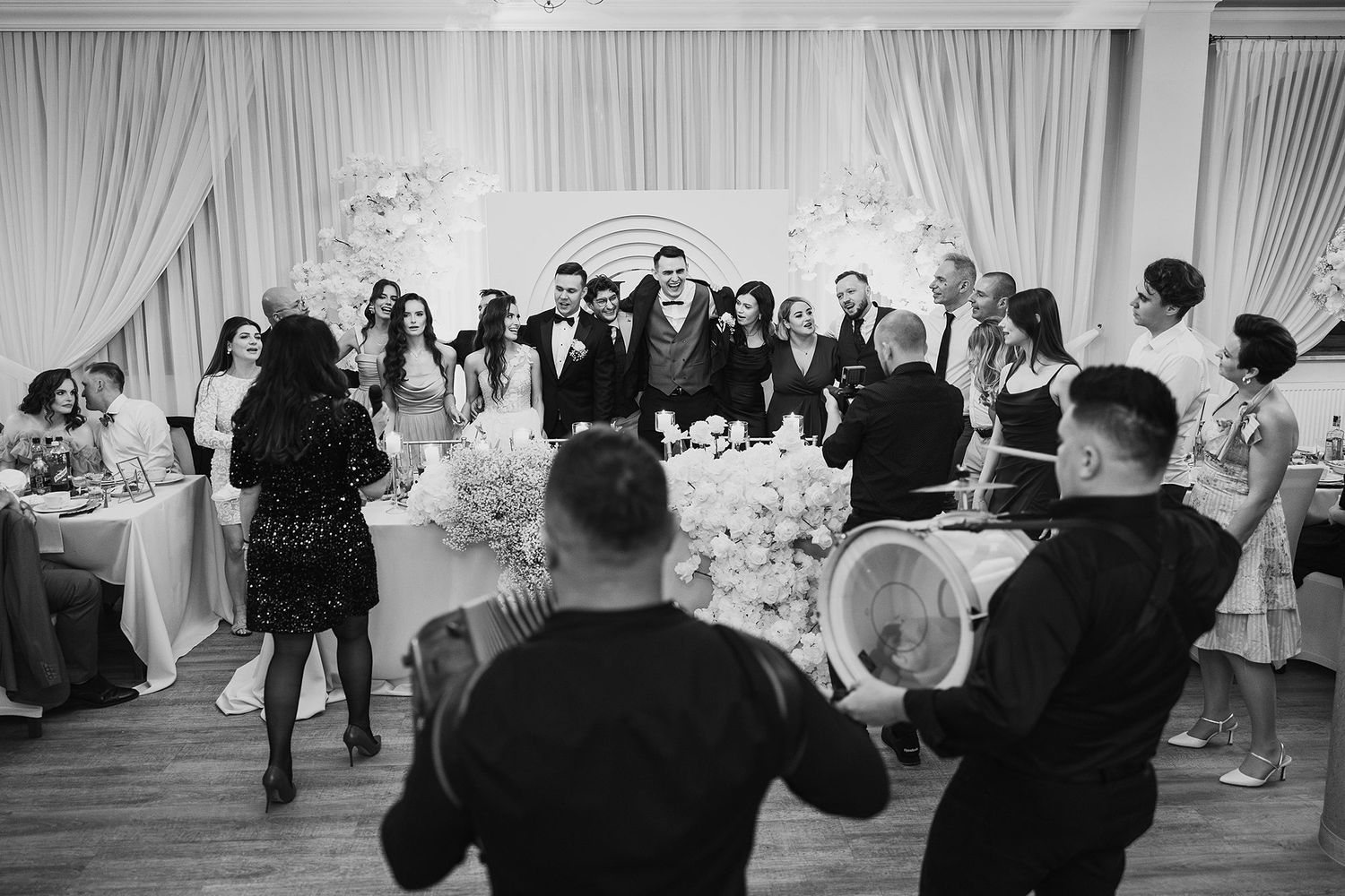 Black and white photo of wedding guests gathered around couple at elegant floral reception venue.