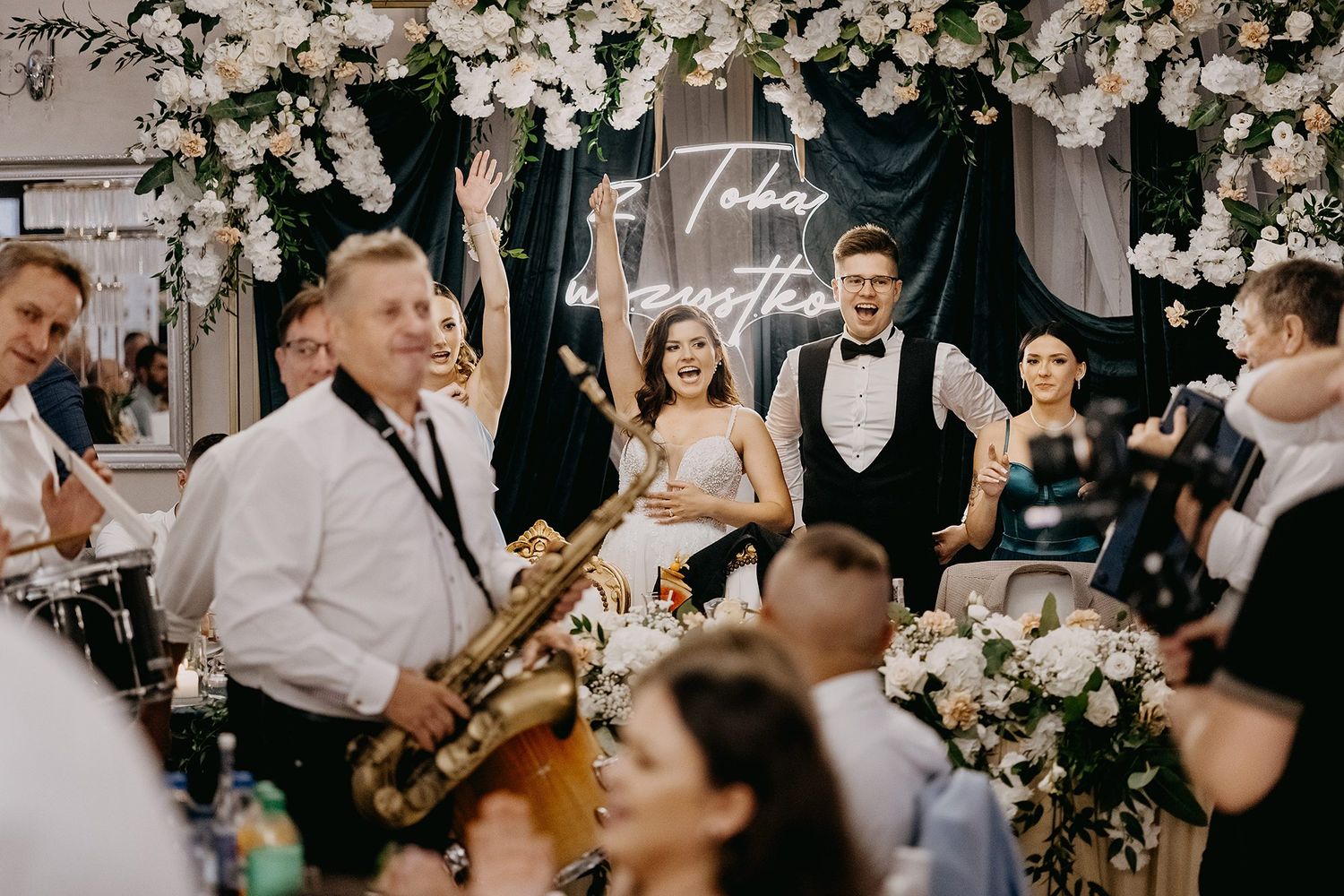 Bride and groom celebrating at wedding reception with live band, saxophone player, and floral decorations.