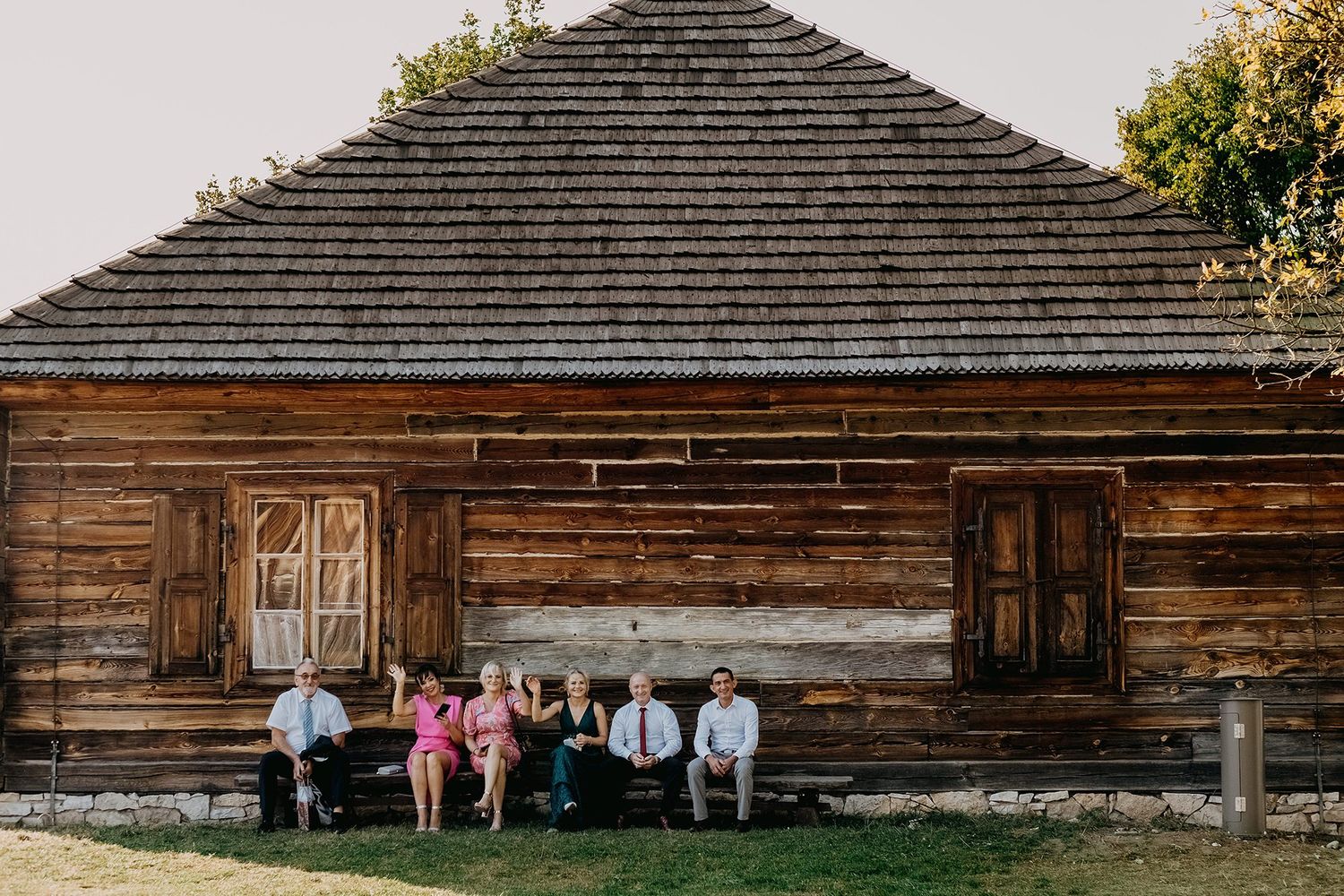 Wedding guests relaxing on bench in front of rustic wooden barn building on sunny day.