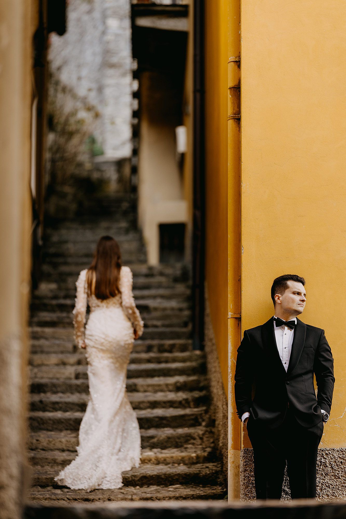 Groom in black tuxedo stands by yellow wall as bride in lace gown ascends cobblestone stairs.