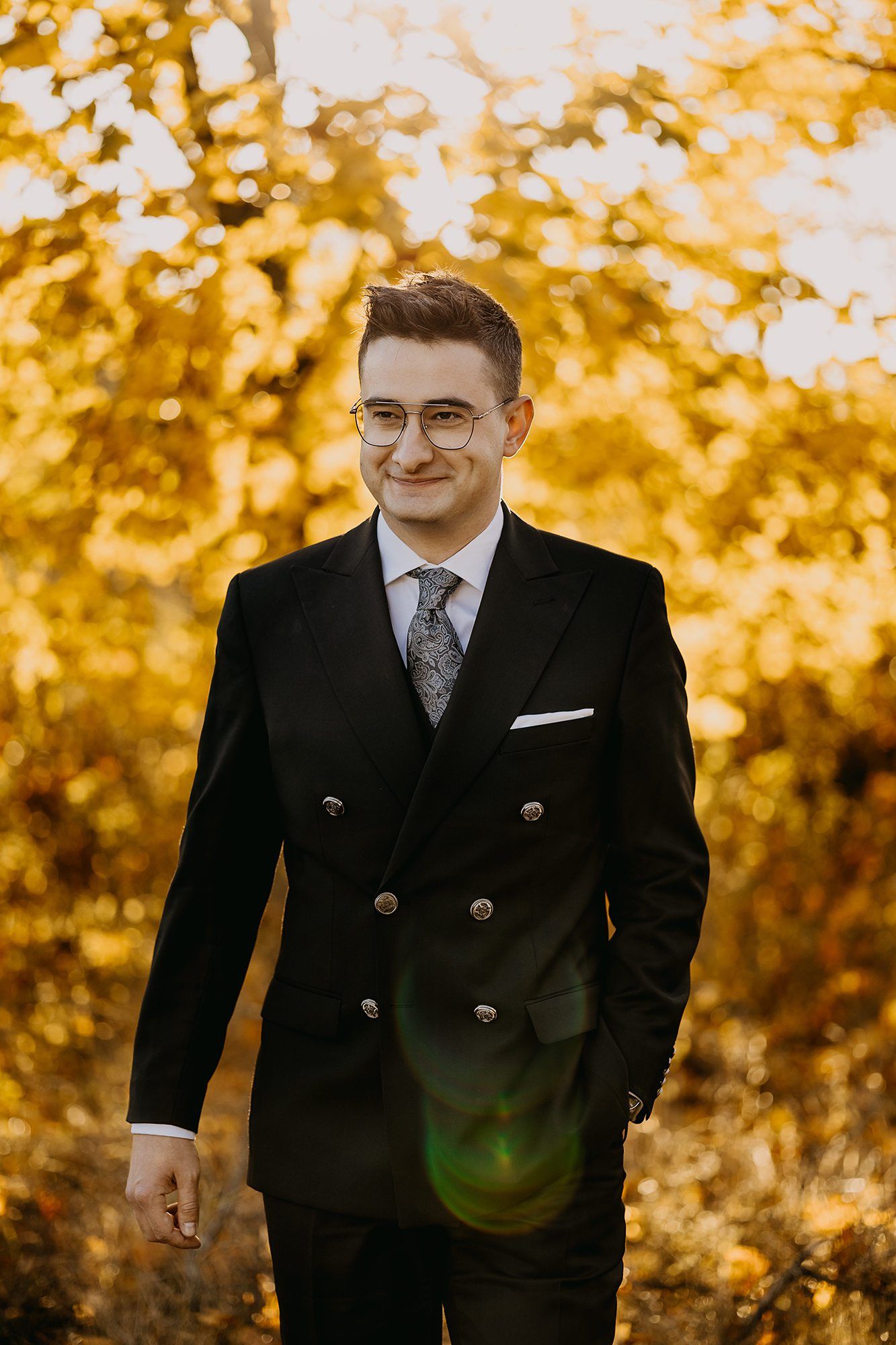 Stylish groom in black double-breasted suit with glasses smiling among vibrant autumn golden foliage.
