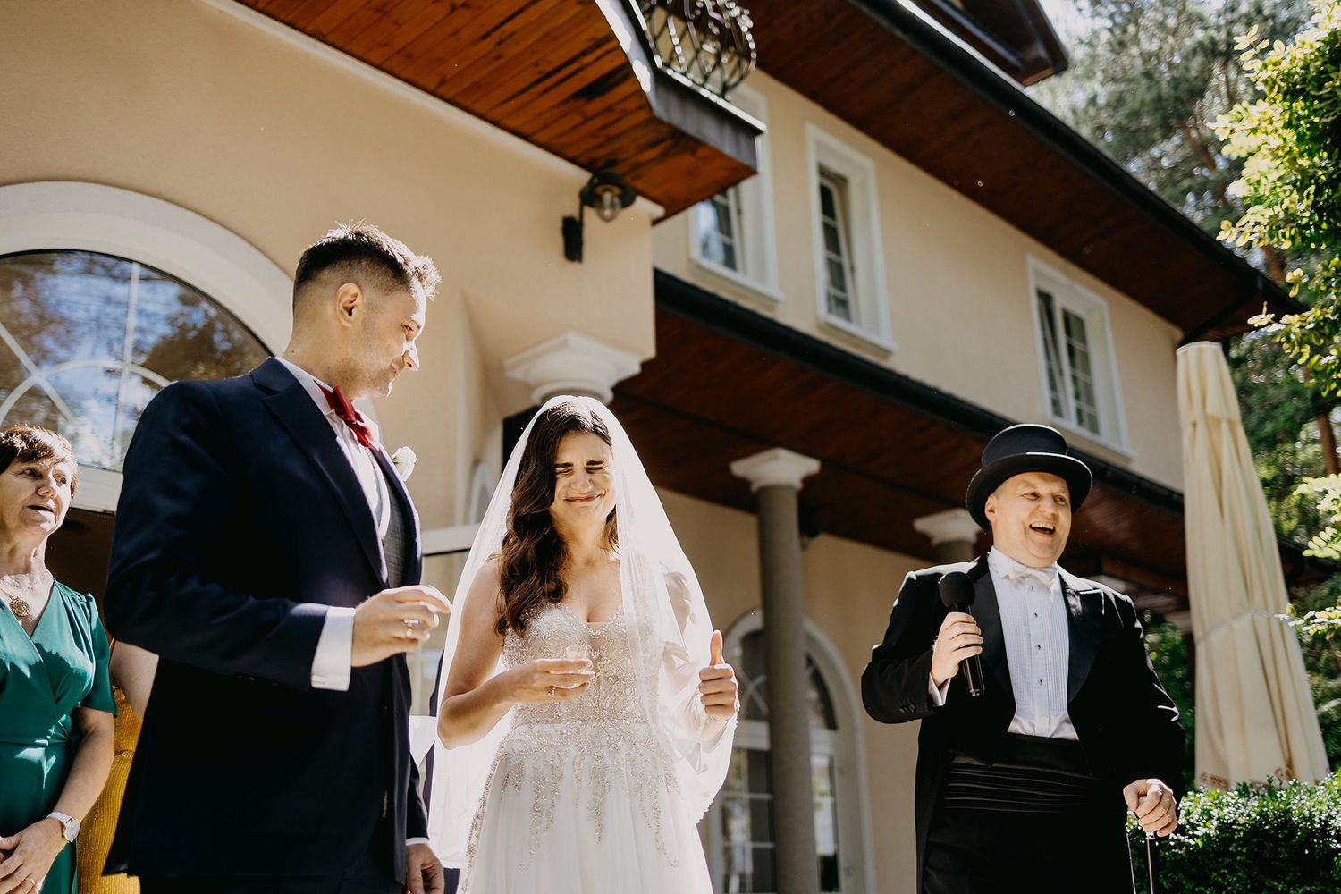 A bride in a white veil and groom stand with guests outside an elegant villa during a wedding reception toast.