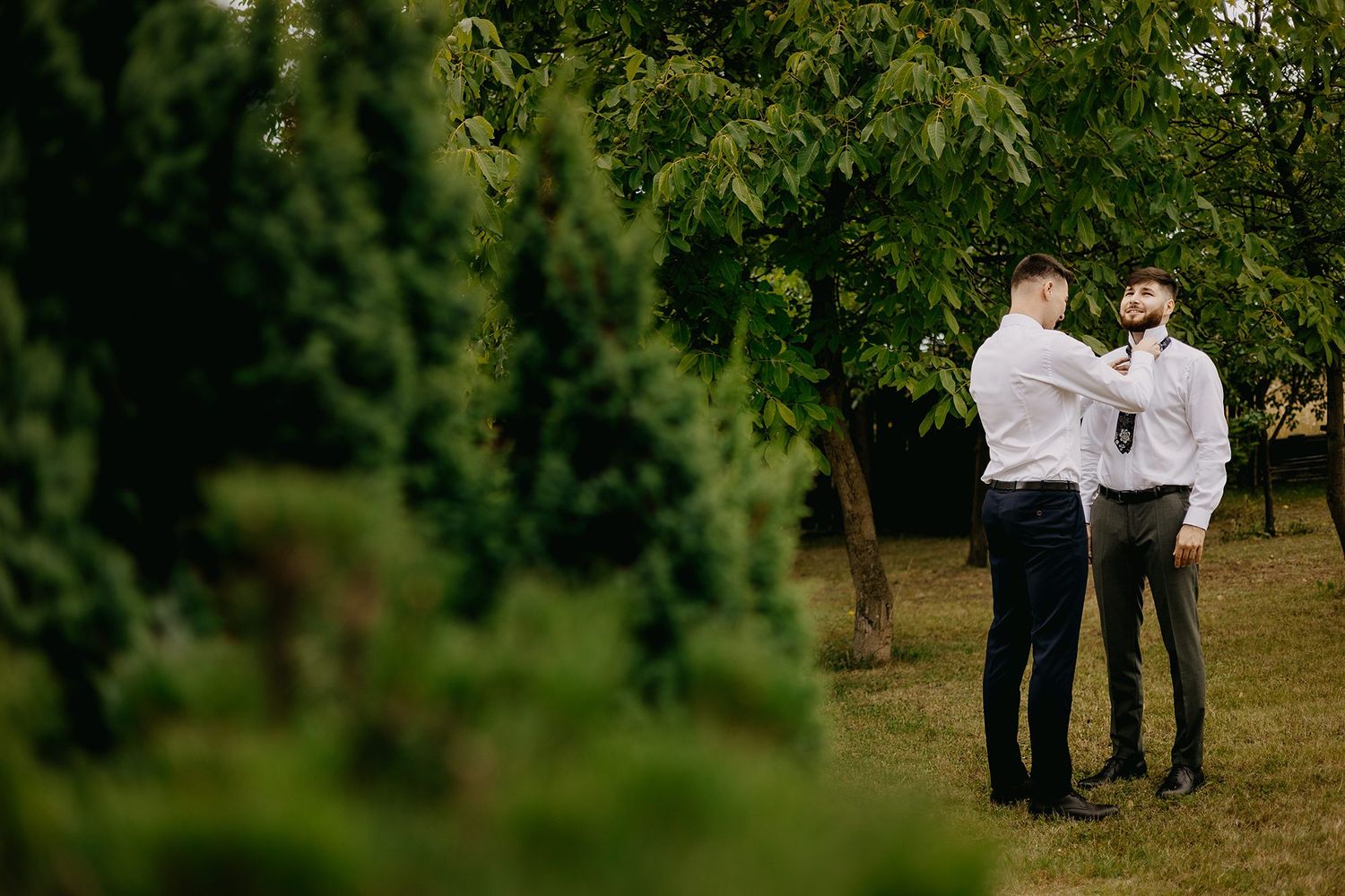Two groomsmen in white shirts adjusting ties in lush green garden before wedding ceremony.