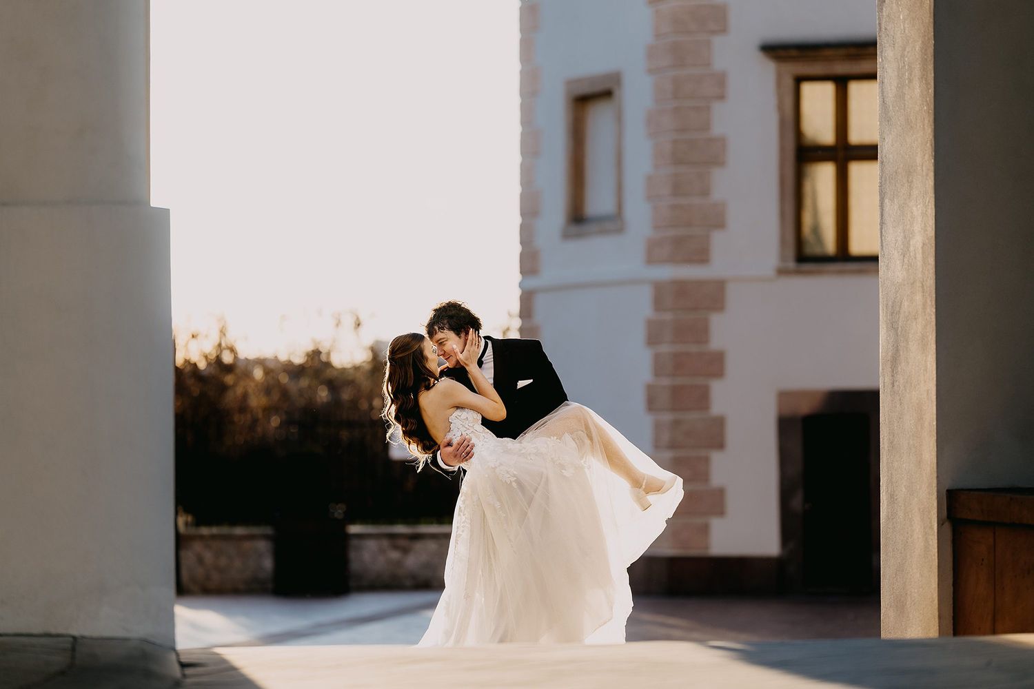 Groom dipping bride in a romantic kiss outdoors in golden sunlight near a white building.