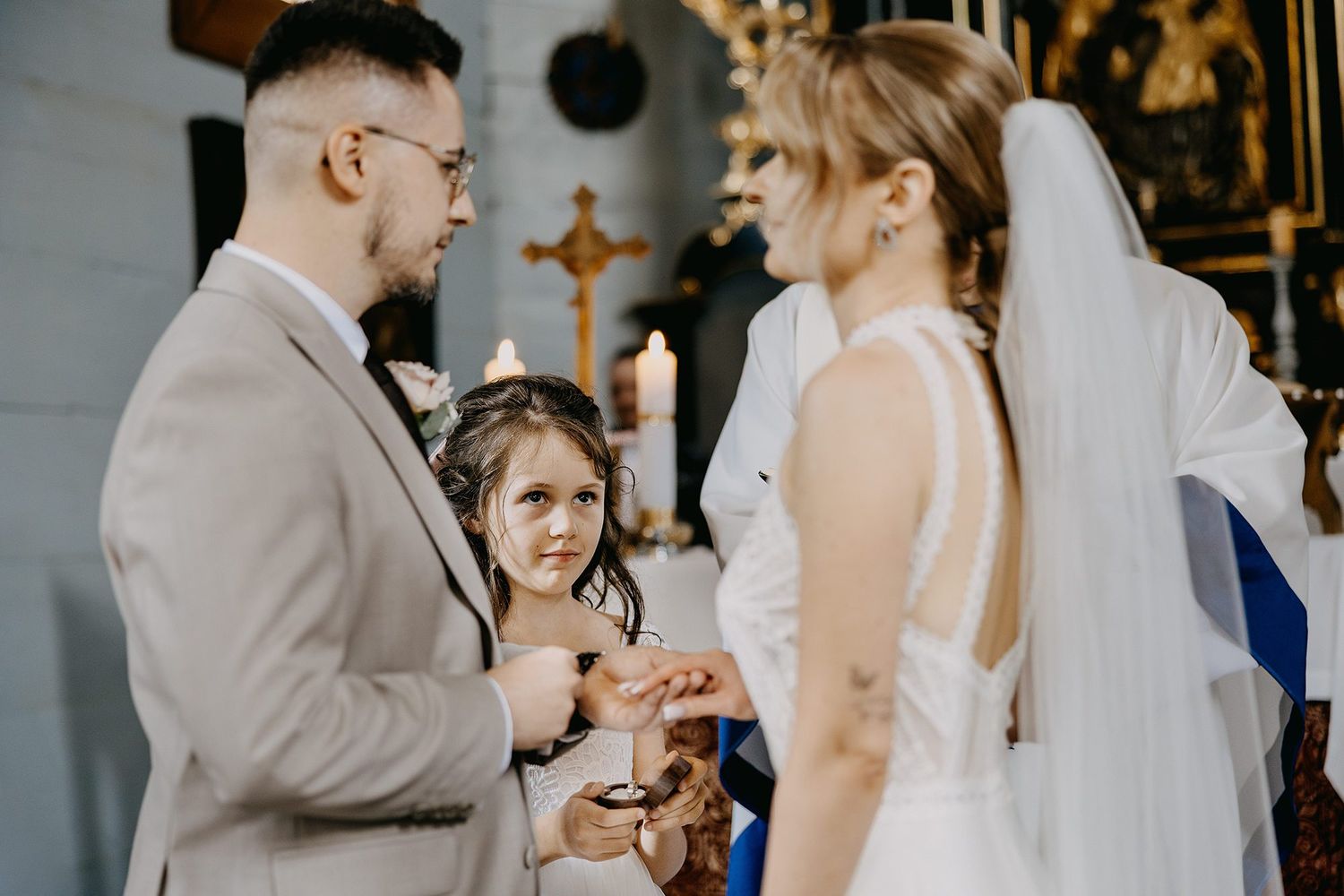 Groom placing wedding ring on bride's finger during church ceremony with young flower girl watching.