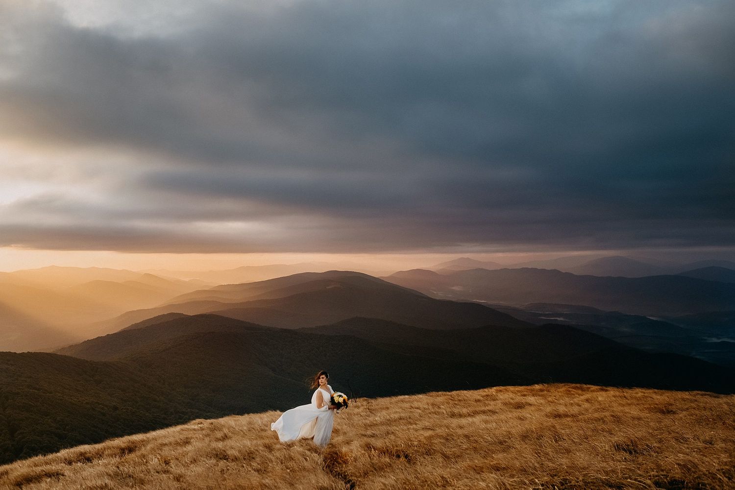 A bride in a flowing white gown stands alone on a dramatic mountain ridge under stormy skies.