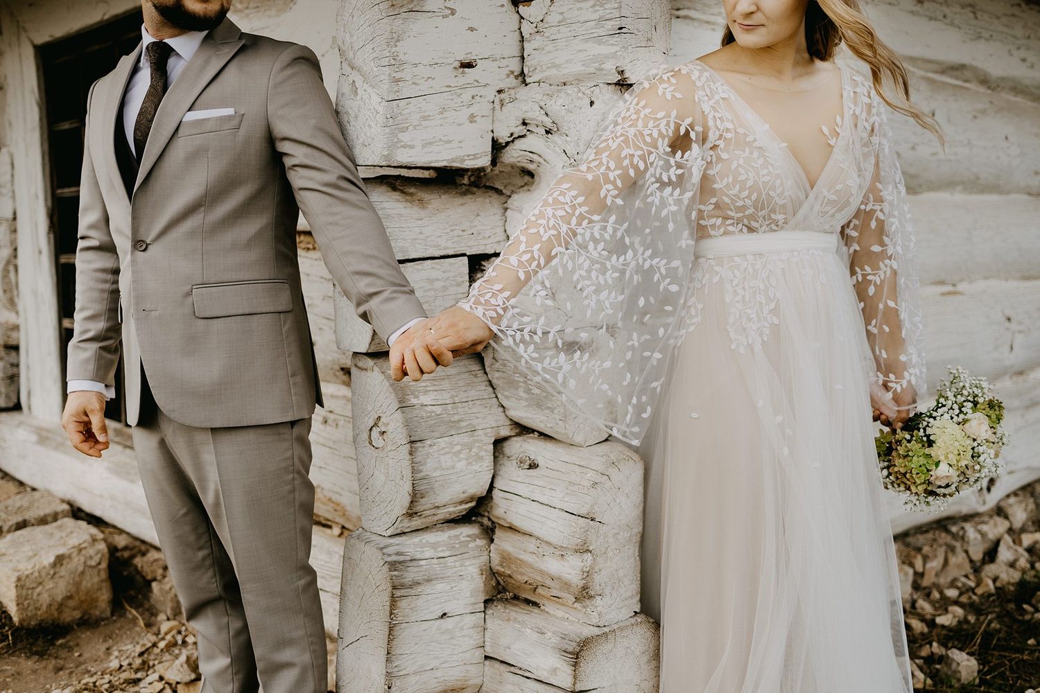 Bride in a pearl-embellished gown and groom in gray suit holding hands near a rustic log wall.