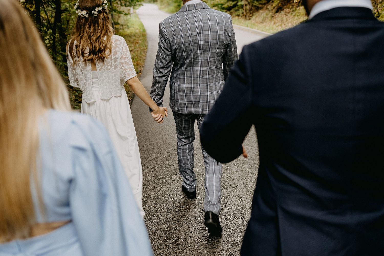 Rear view of bride and groom in plaid suit walking hand in hand down a tree-lined path.