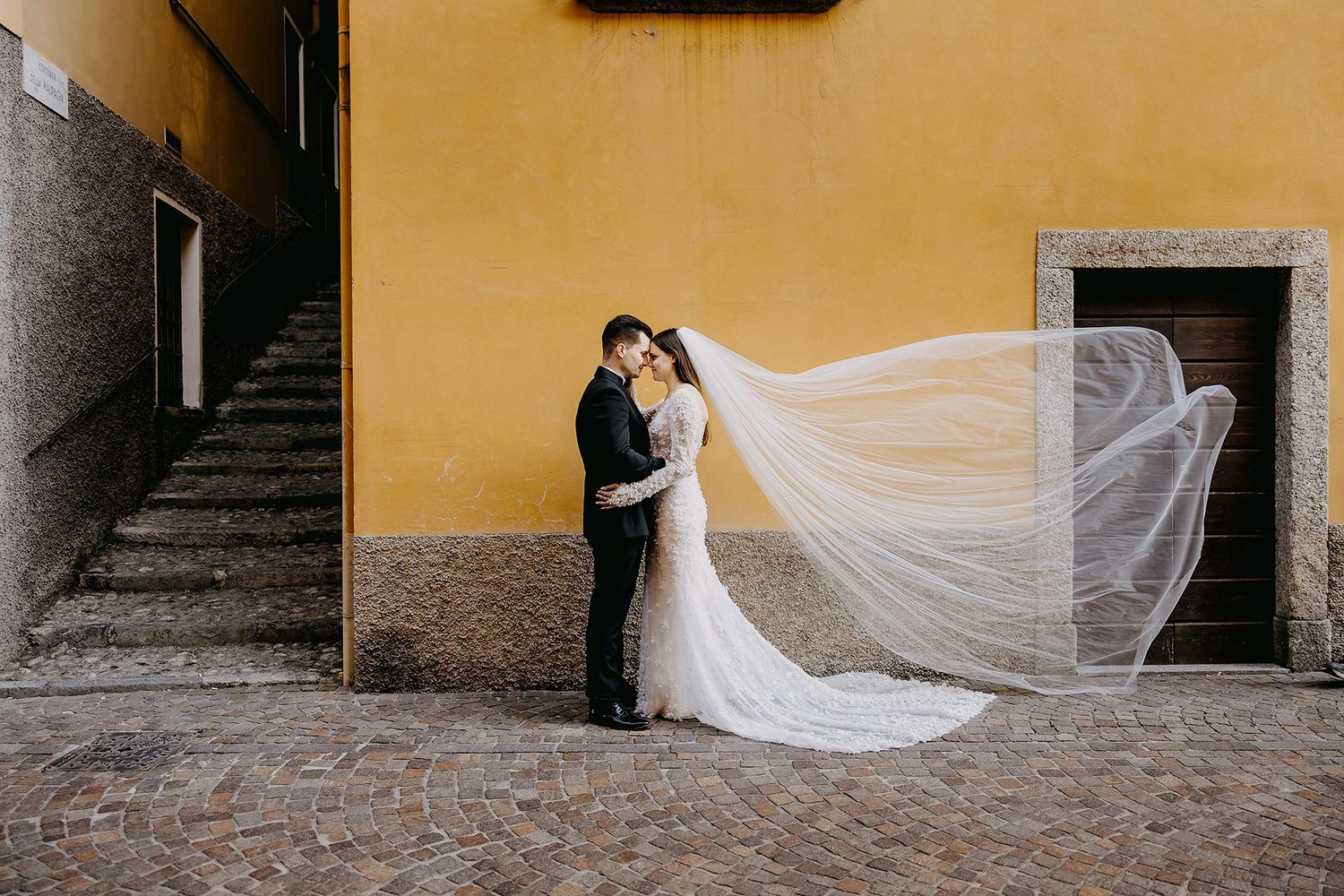 Wedding couple embraces as bride's long veil billows dramatically against a yellow Italian wall.