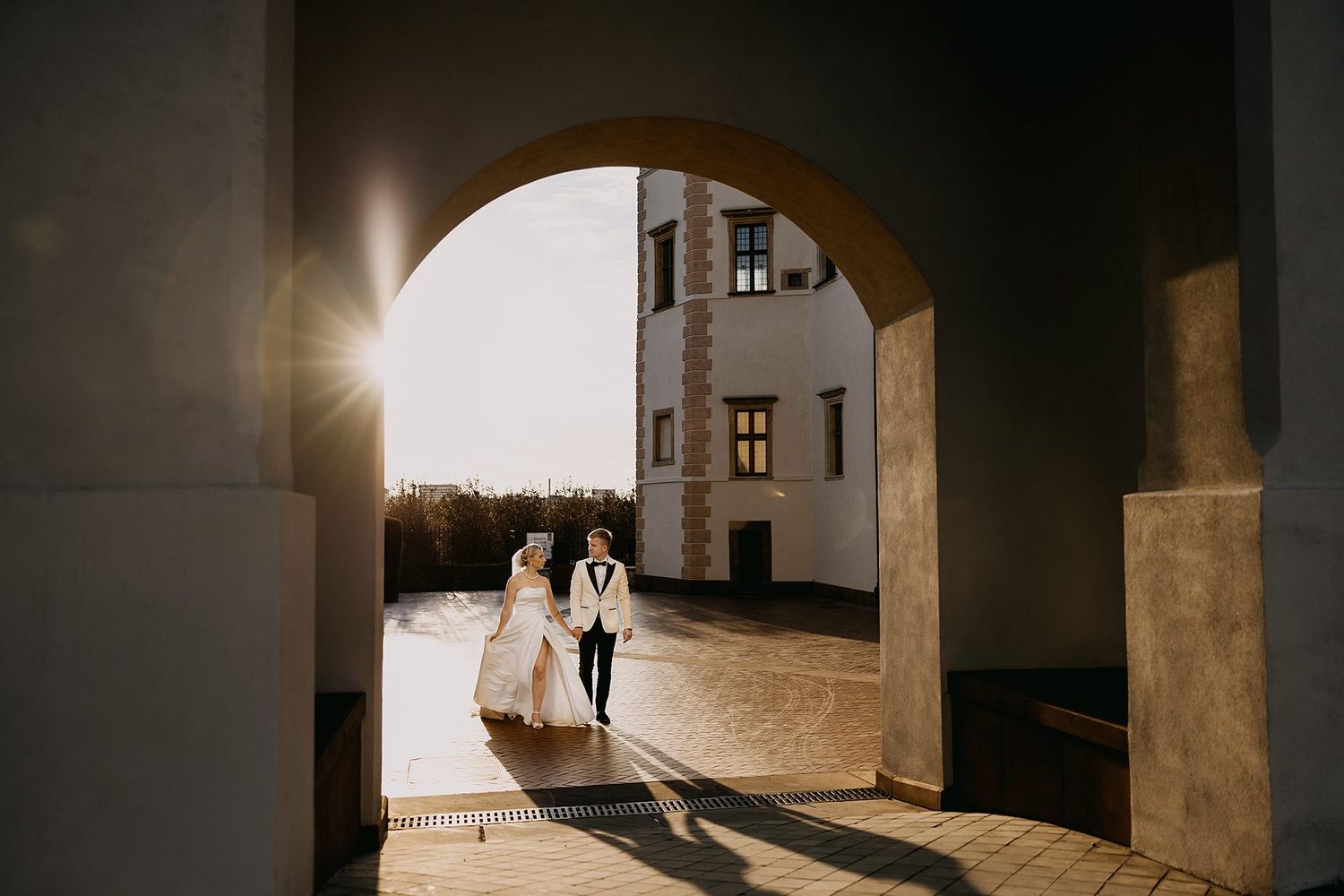 Bride and groom walking hand in hand through a sunlit archway in a historic courtyard venue.