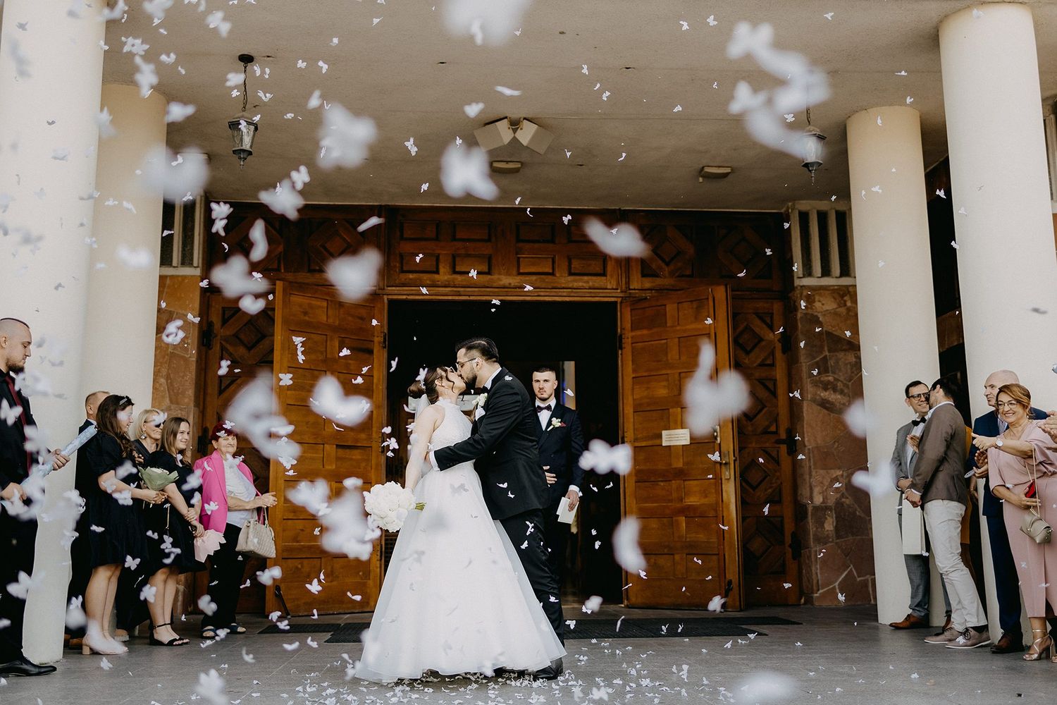Newlyweds kiss in shower of white confetti petals outside church surrounded by wedding guests.