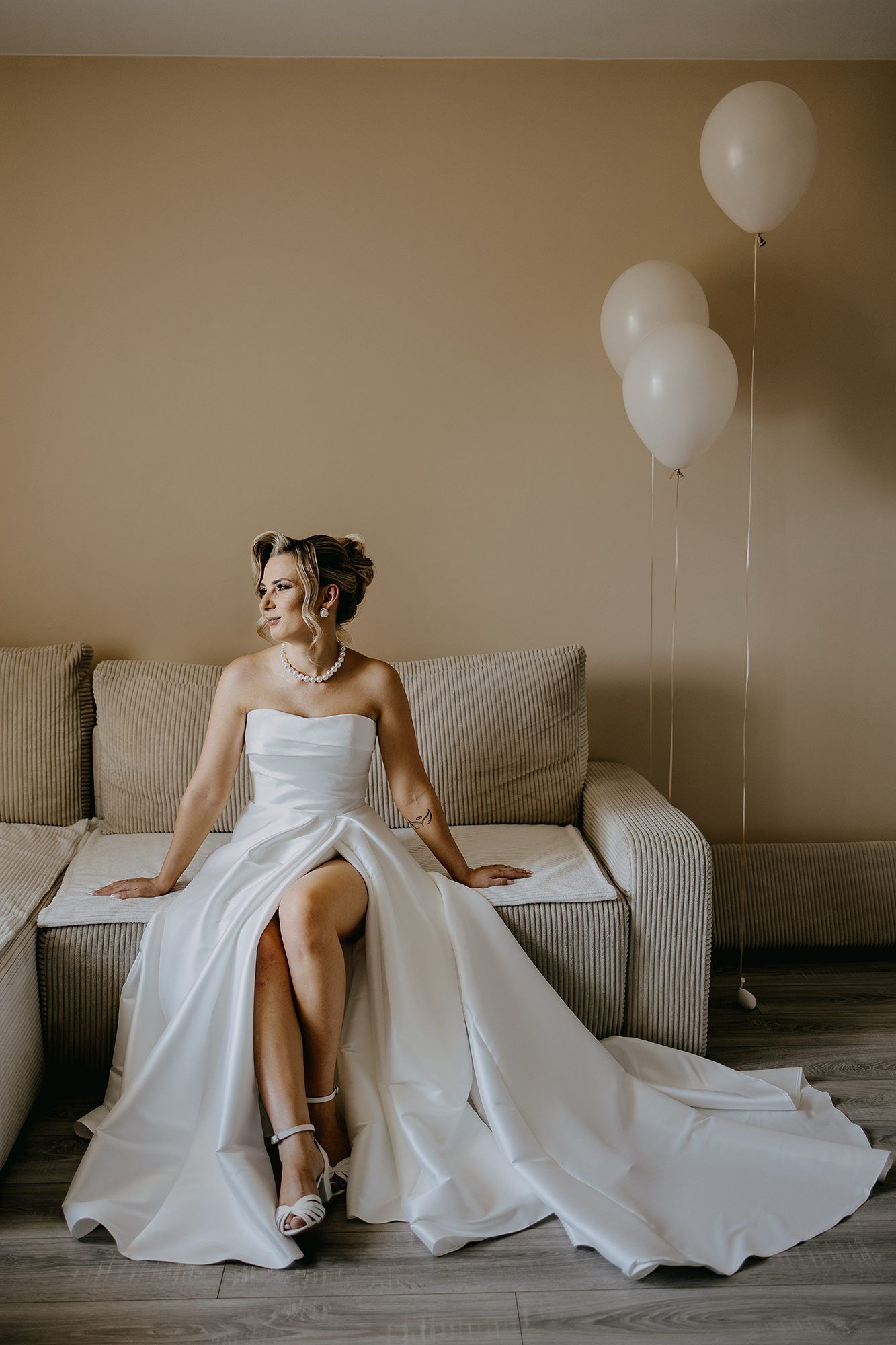 Bride in elegant strapless white gown with slit seated on sofa next to two white balloons.