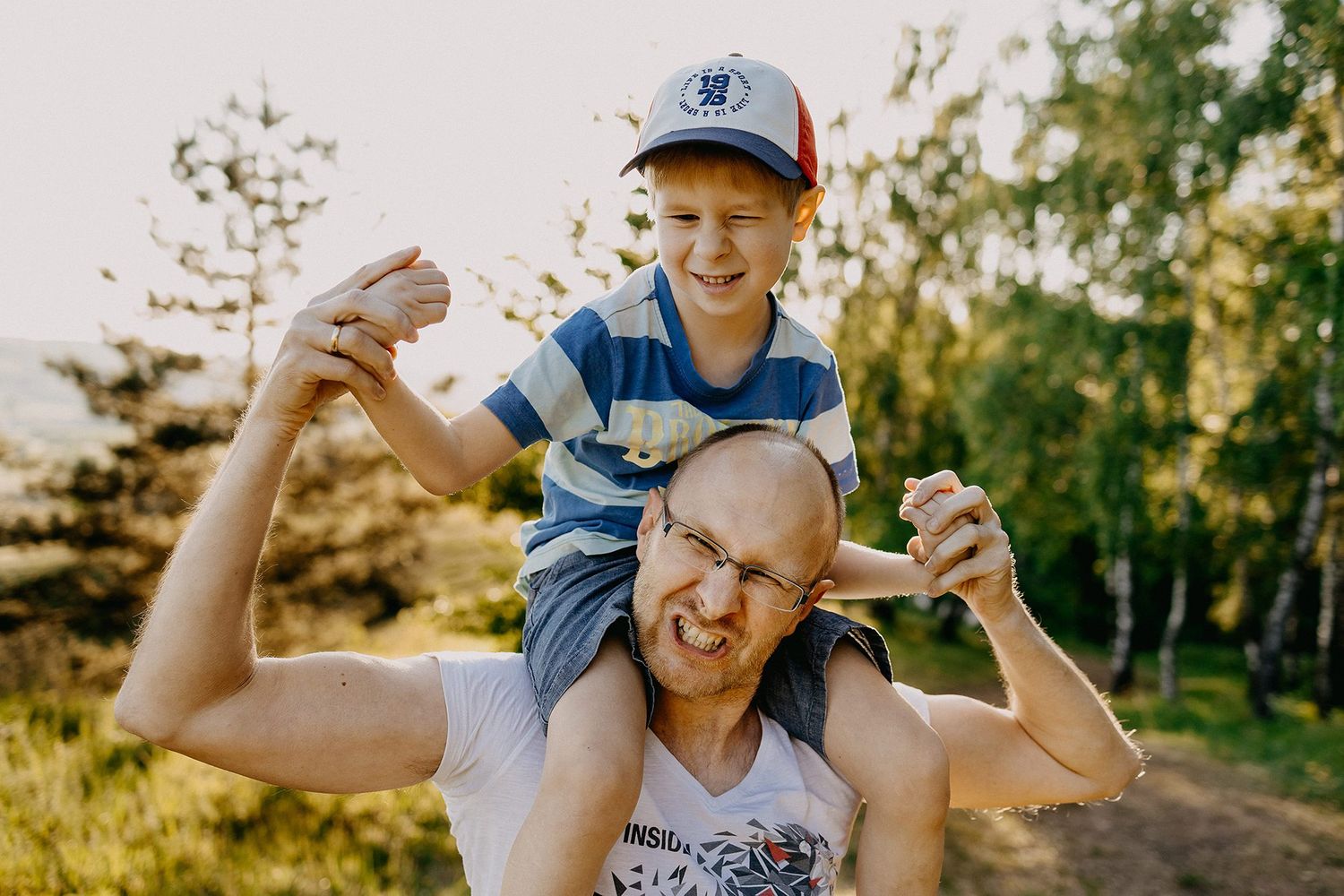 Boy sitting on fathers shoulders outdoors in sunlight.