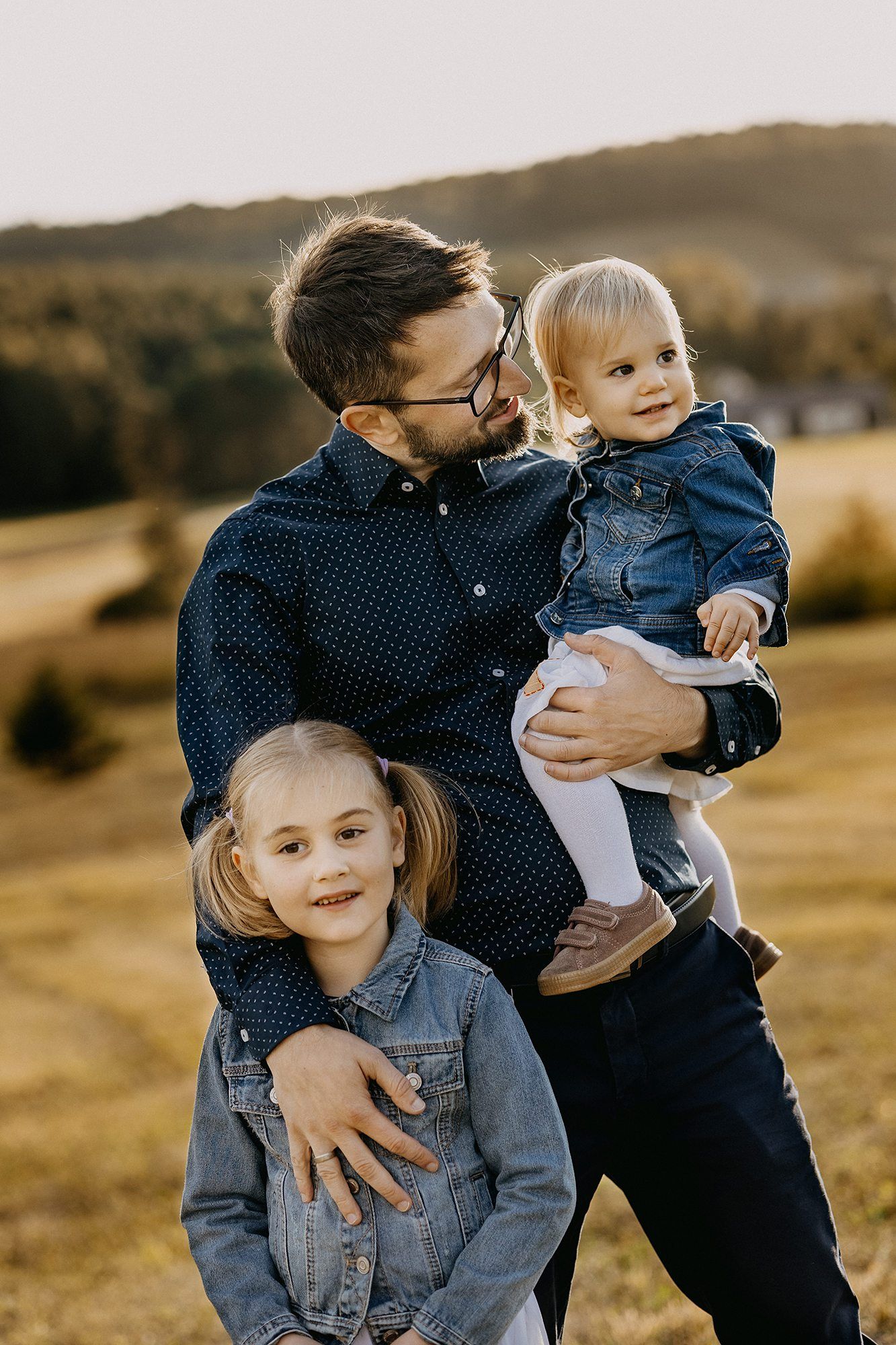 Father holding toddler while hugging older daughter in sunny autumn field.