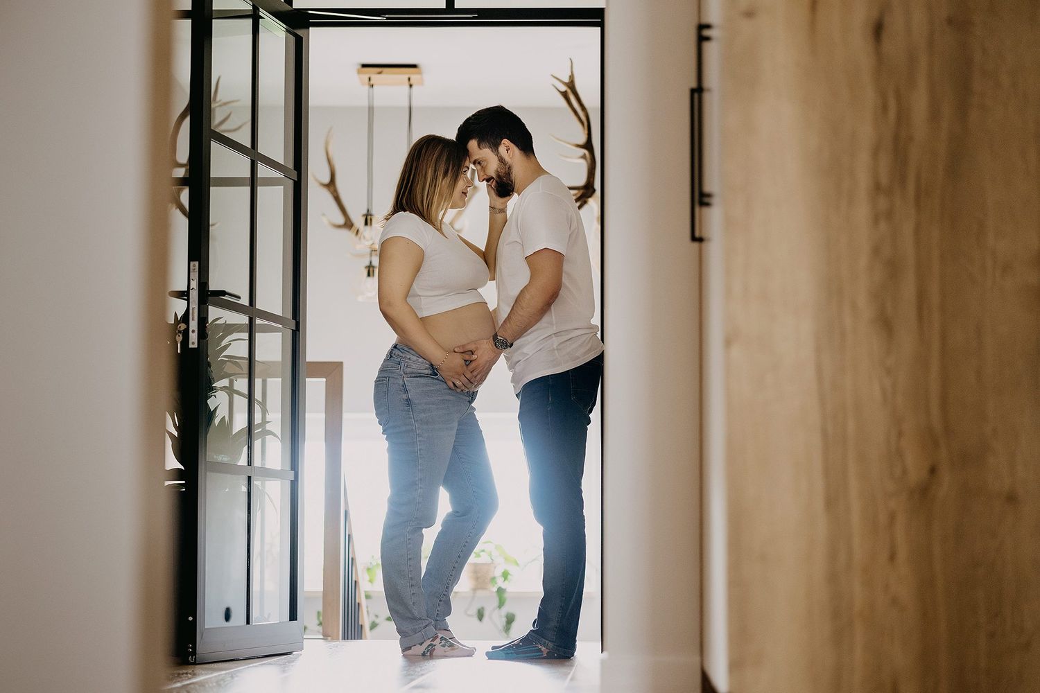 Pregnant couple sharing a tender moment in a sunlit doorway, touching foreheads and holding baby bump.