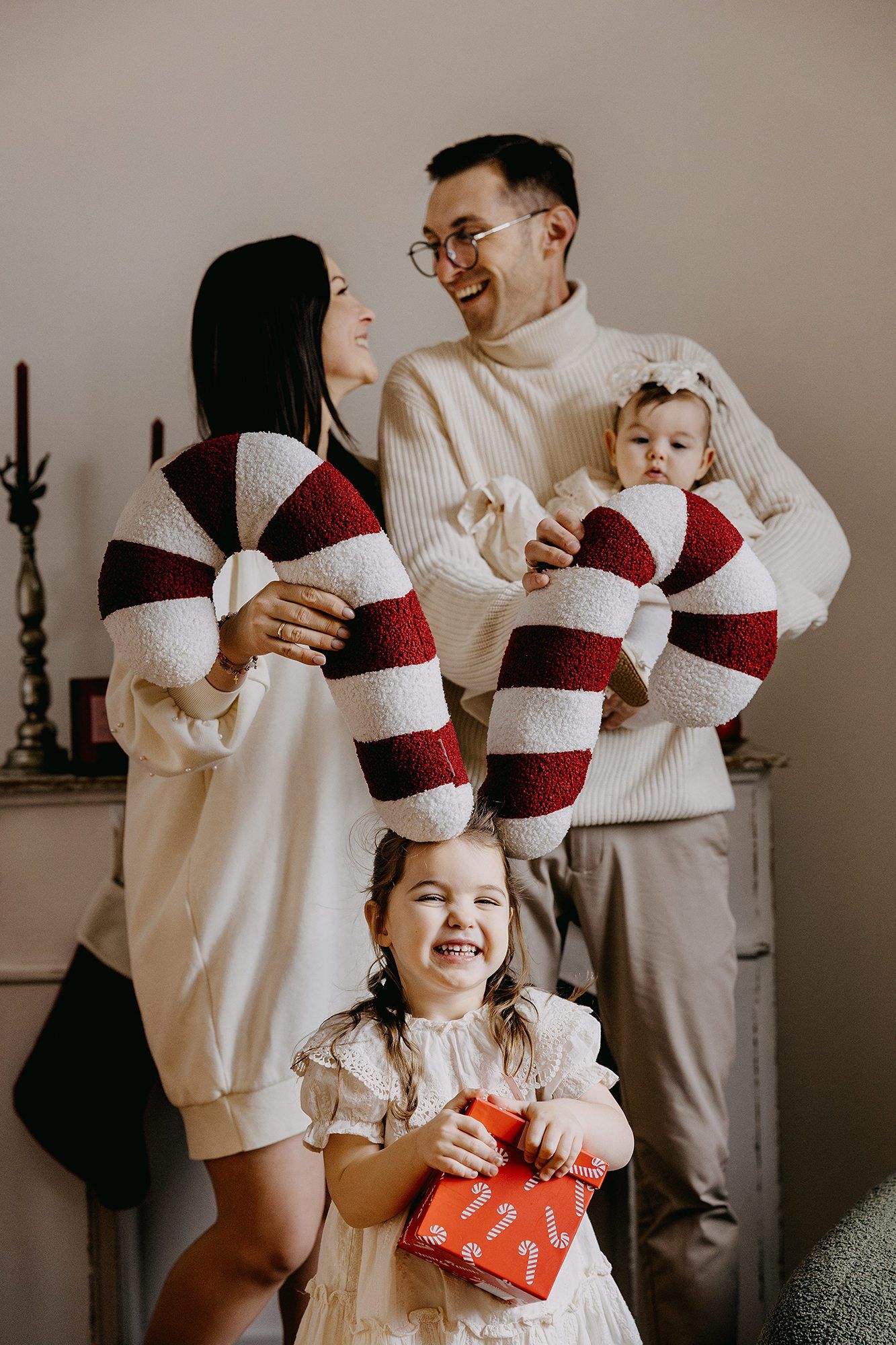 A joyful family poses with large candy cane decorations while their daughter laughs holding a wrapped Christmas gift.