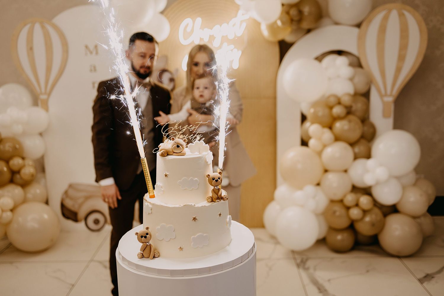 A couple with child celebrates a christening with a two-tier white cake and sparklers.