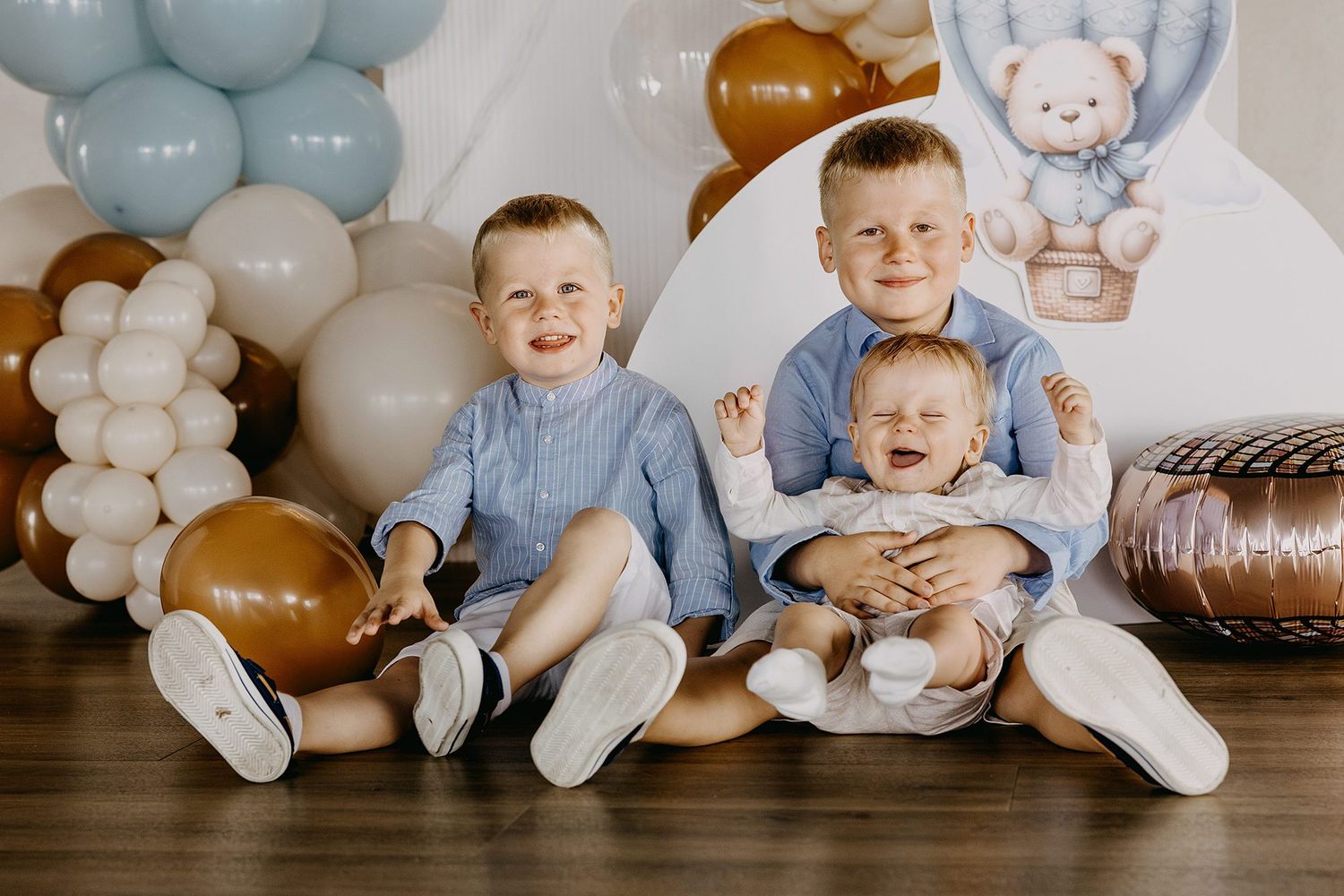 Three young brothers posing together with balloons and teddy bear decorations.