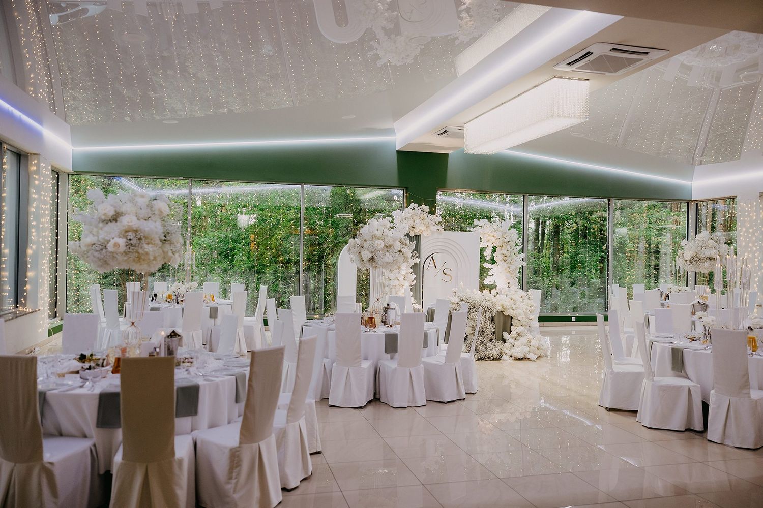 Elegant wedding reception hall with white chair covers, floral arrangements, and floor-to-ceiling windows overlooking lush greenery.