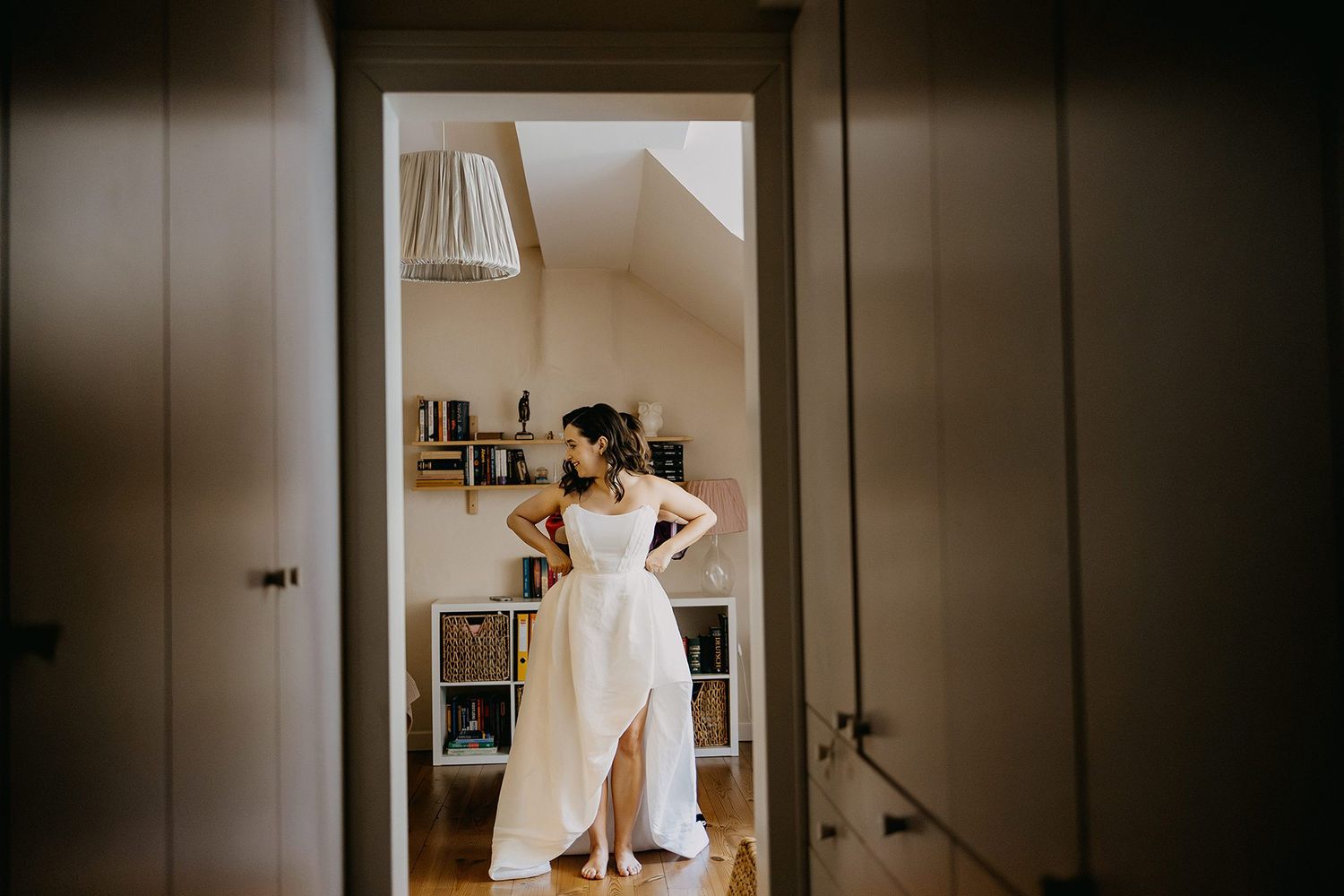 Bride in white wedding dress with slit stands confidently in a sunlit room, hands on hips, surrounded by bookshelves.