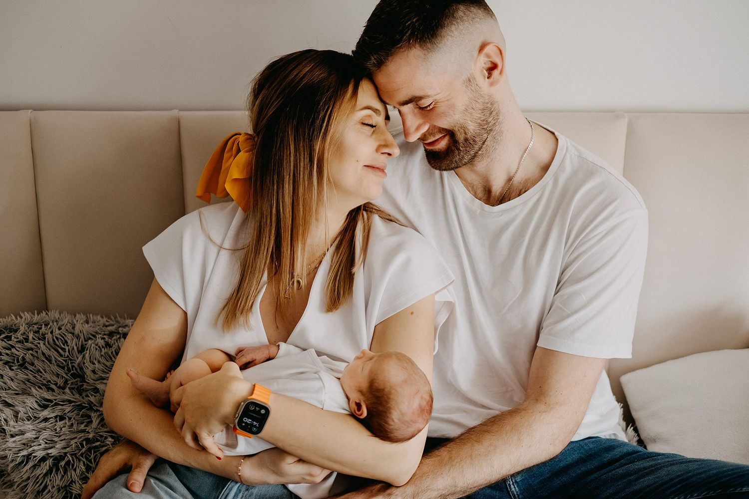 A smiling couple sits on a bed, lovingly holding their newborn baby together.