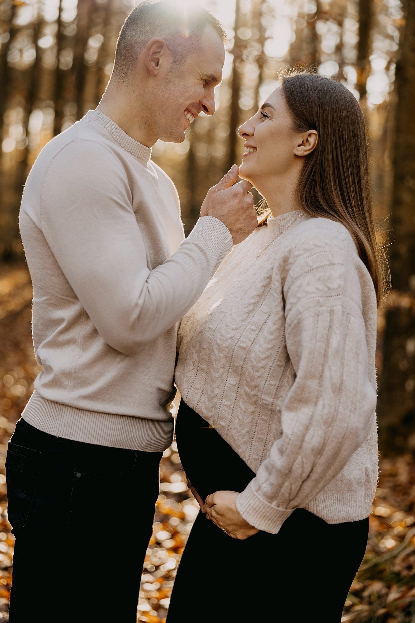 Smiling couple sharing a tender moment outdoors in a sunlit autumn forest during maternity session.