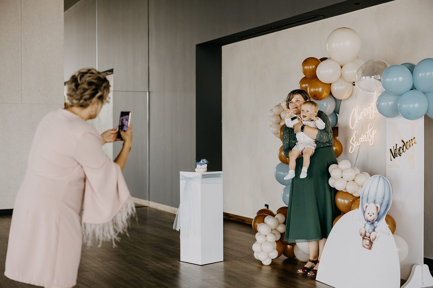 Woman photographing a mother holding her baby at a christening balloon backdrop celebration.