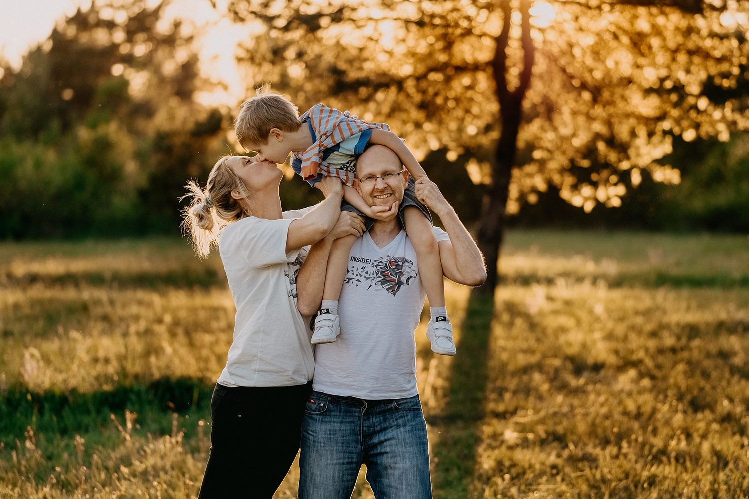 Playful family moment outdoors at sunset with child sitting on fathers shoulders while mother kisses him.