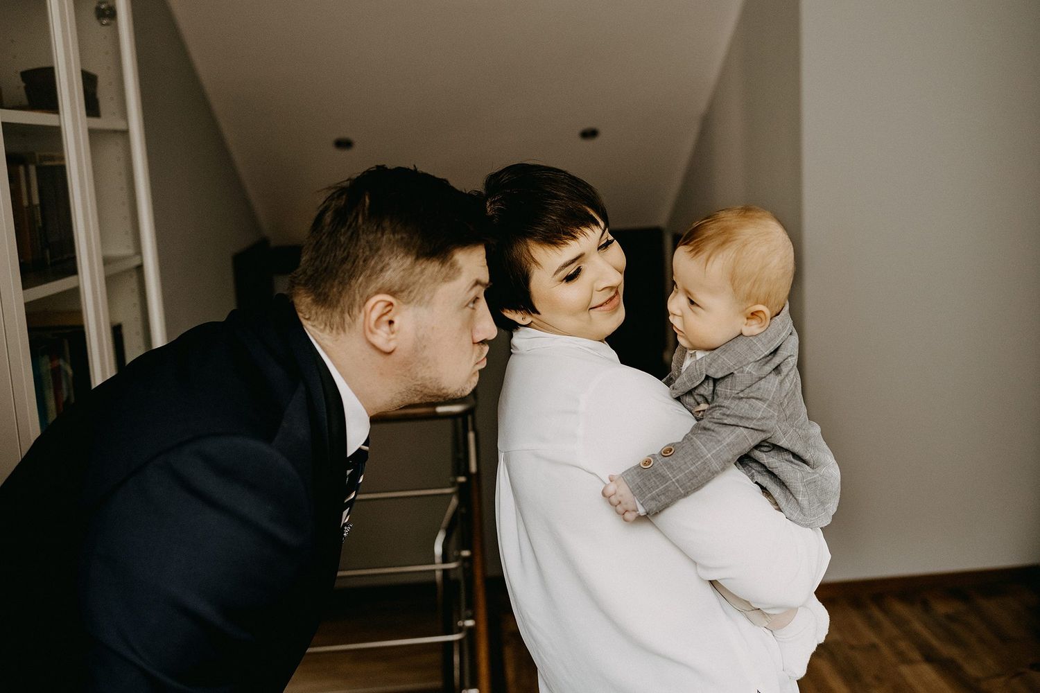 A couple shares a tender moment with their baby, the father leaning in for a kiss in a cozy home setting.