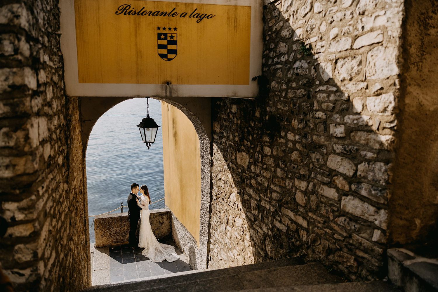 Wedding couple kisses under a stone archway with a glimpse of the lake and Italian restaurant sign.