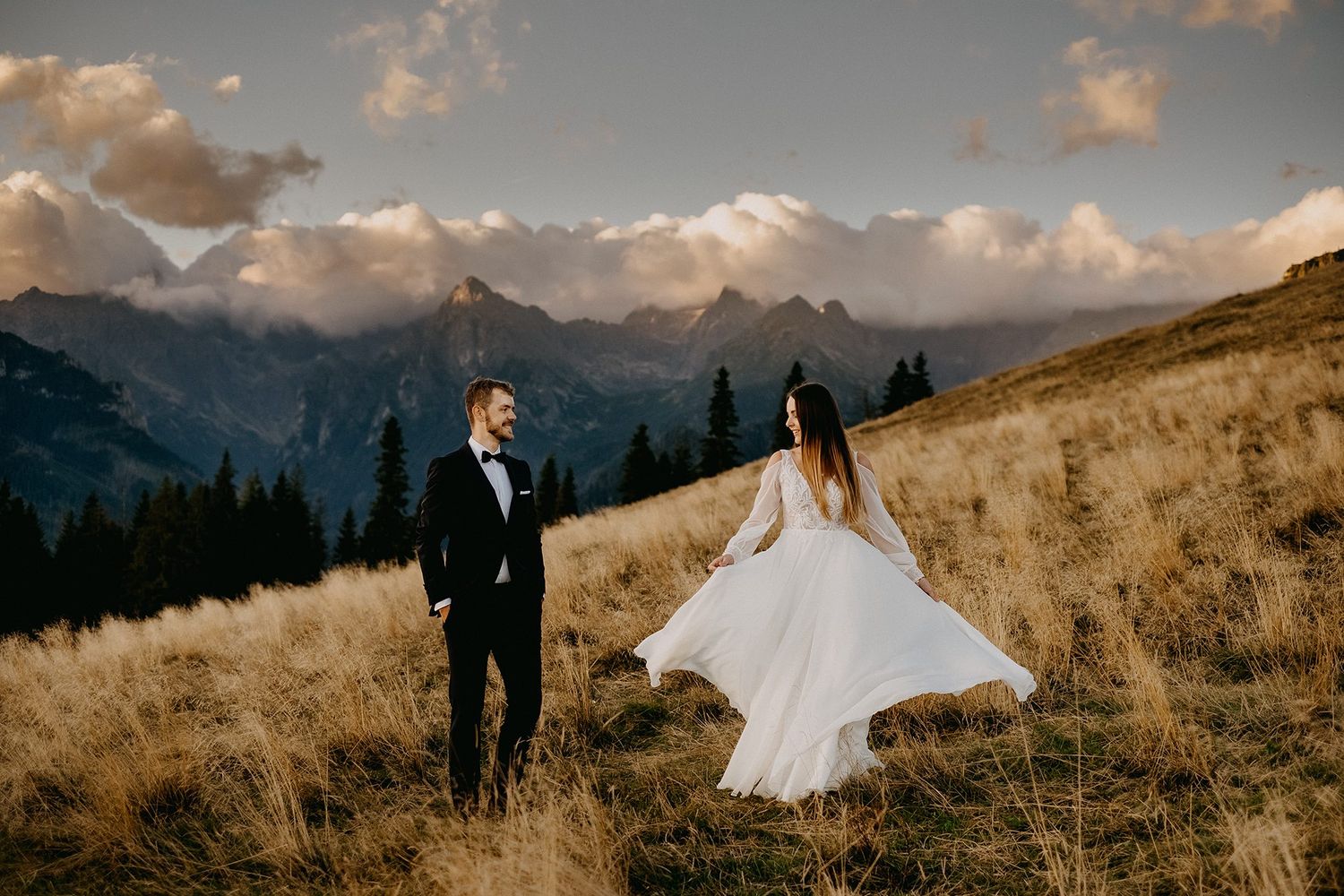Bride in flowing white gown and groom in black tux walk through golden mountain meadow at sunset.