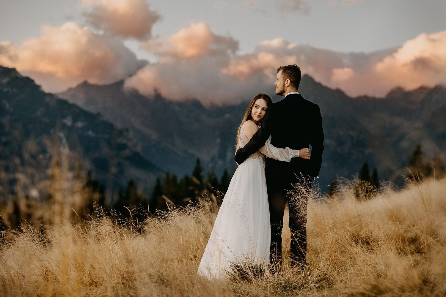 Bride and groom share a romantic embrace in a golden meadow with dramatic mountain backdrop.