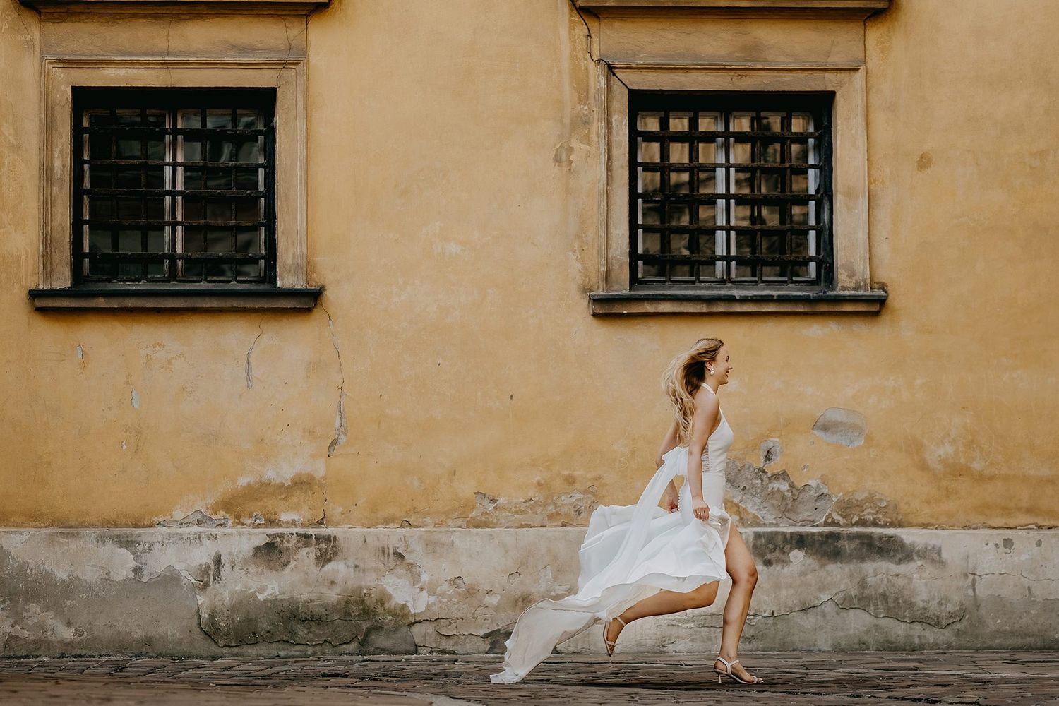 Bride in flowing white gown runs joyfully along a weathered yellow stone wall with barred windows.