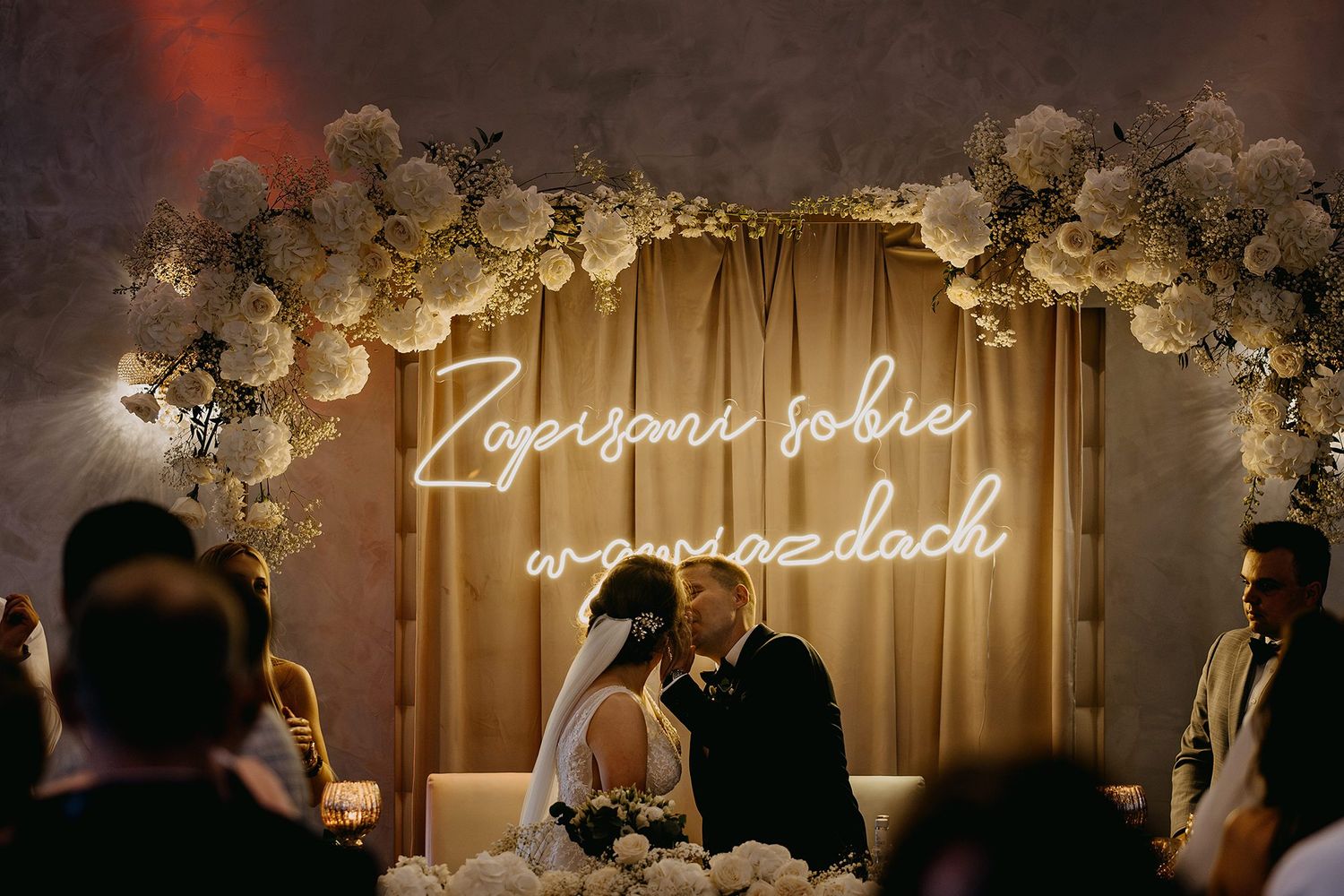 Couple kissing under neon sign and white floral arch at wedding reception.