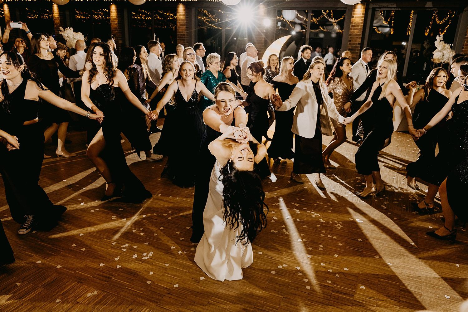 Energetic wedding reception dance floor filled with guests in black dresses surrounding the dancing bride.