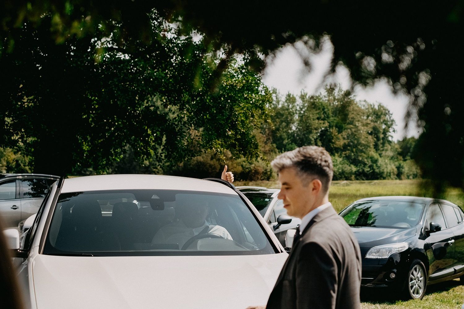Groomsman in suit standing near white wedding car outdoors surrounded by trees.