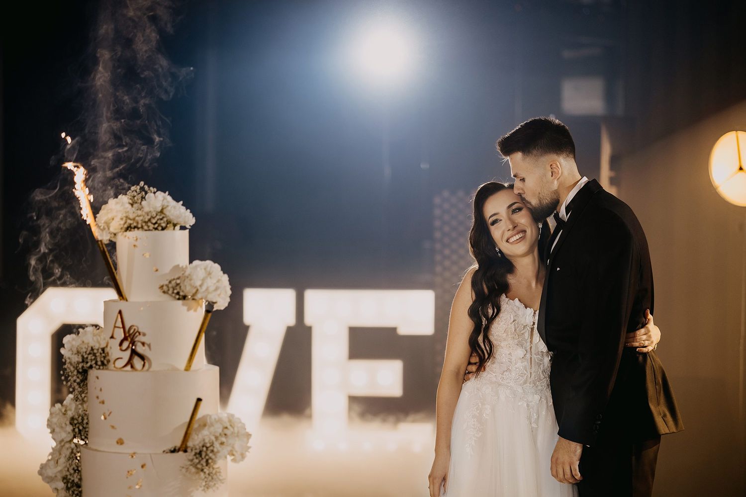 Smiling bride and groom standing beside a white multi-tiered wedding cake with sparklers and a LOVE sign backdrop.