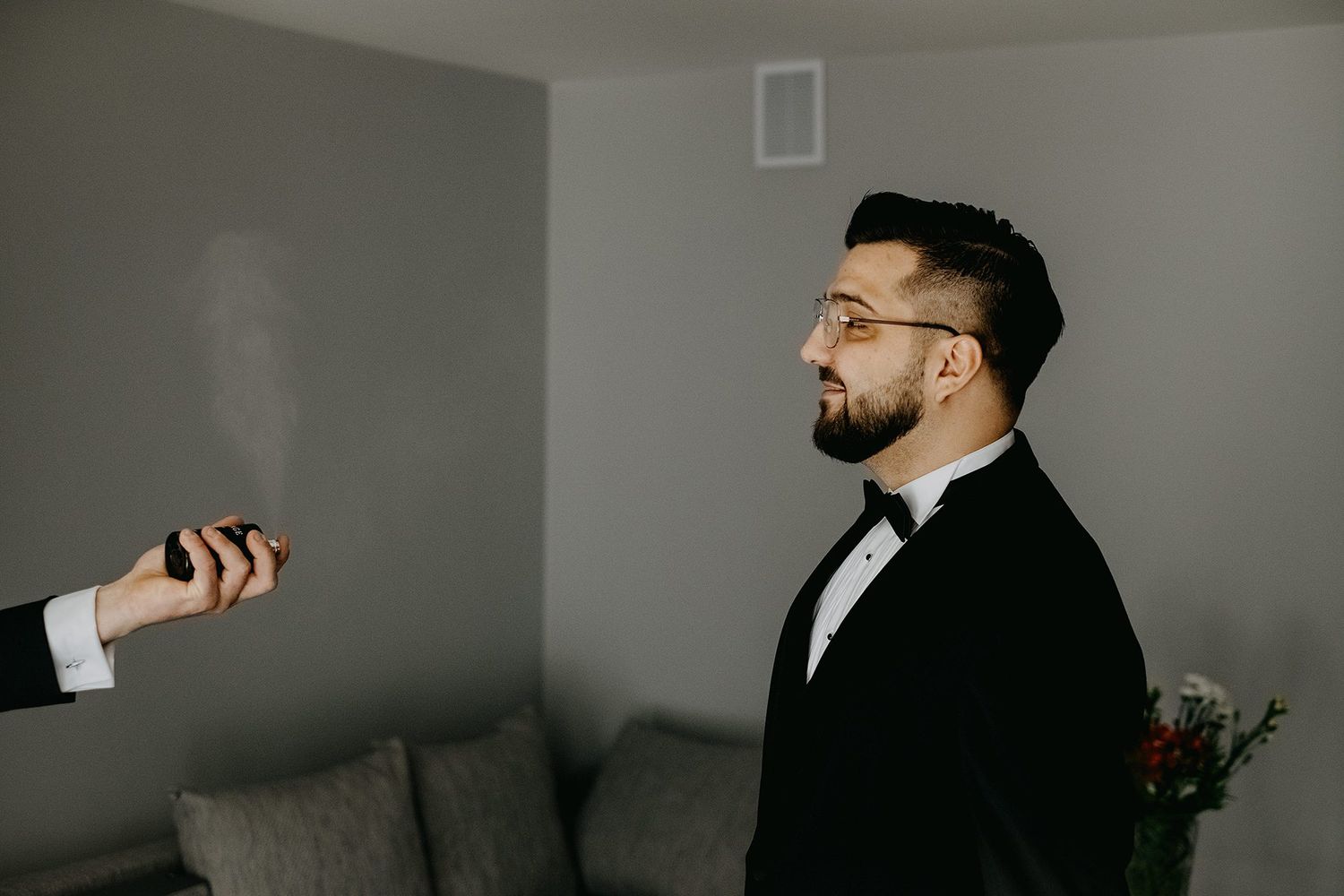 Groom in black tuxedo and bow tie being spritzed with cologne during wedding preparations.