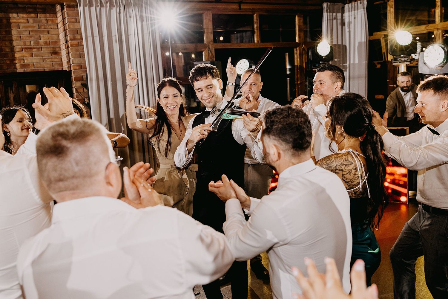 Violinist performing energetically among cheering wedding guests in a warmly lit rustic reception venue.