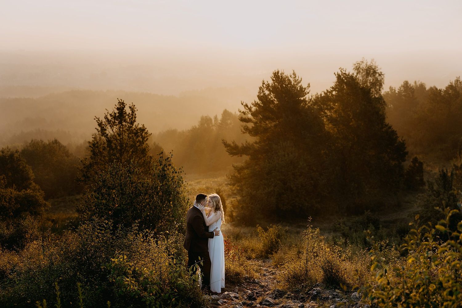 Bride and groom embracing at golden hour in a misty forest landscape with warm light.