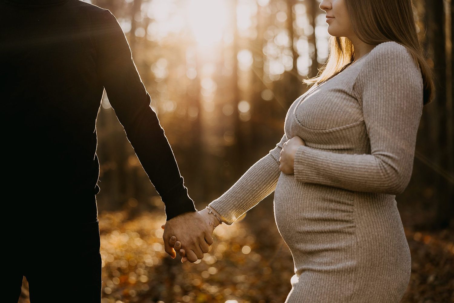 Pregnant woman holds her baby bump while holding hands with partner in golden autumn forest.