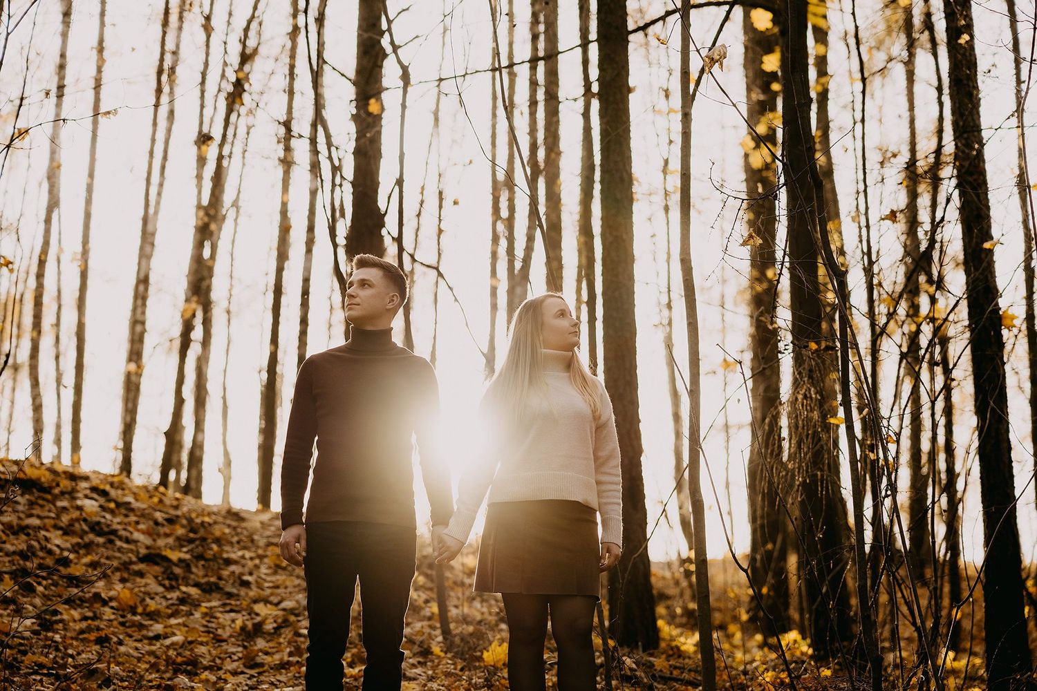Couple holding hands in a sunlit autumn forest with golden leaves and glowing backlight.