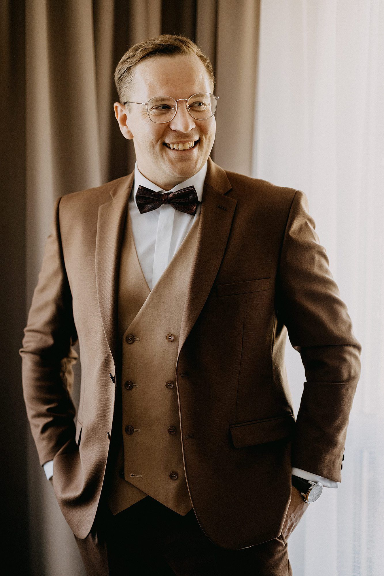 Smiling groom in brown tuxedo with bow tie and glasses posing by window.