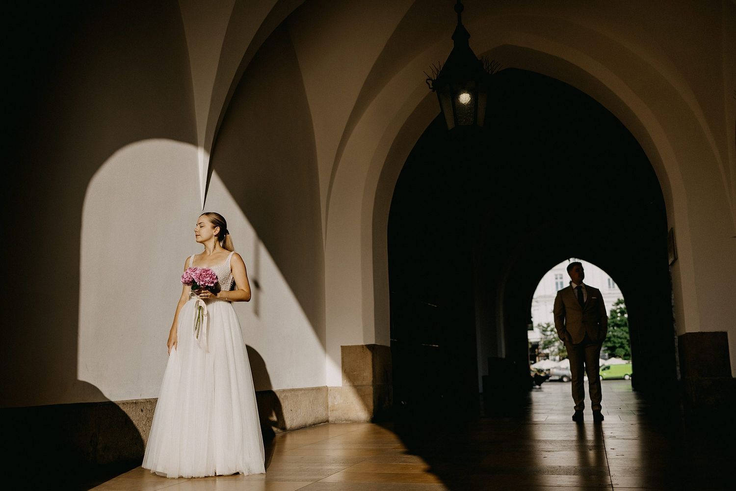 Bride holding a bouquet stands in dramatic arched hallway light while groom walks in the background.